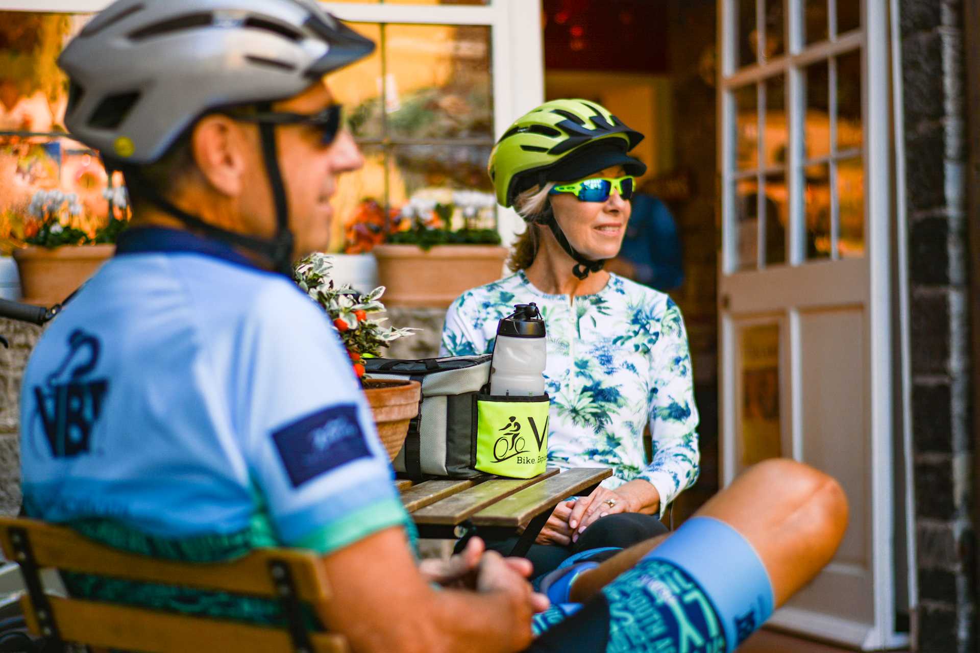 Two cyclists, one wearing a blue jersey and the other a floral-patterned shirt, are conversing in what appears to be a cozy outdoor setting with a wooden structure in the background.