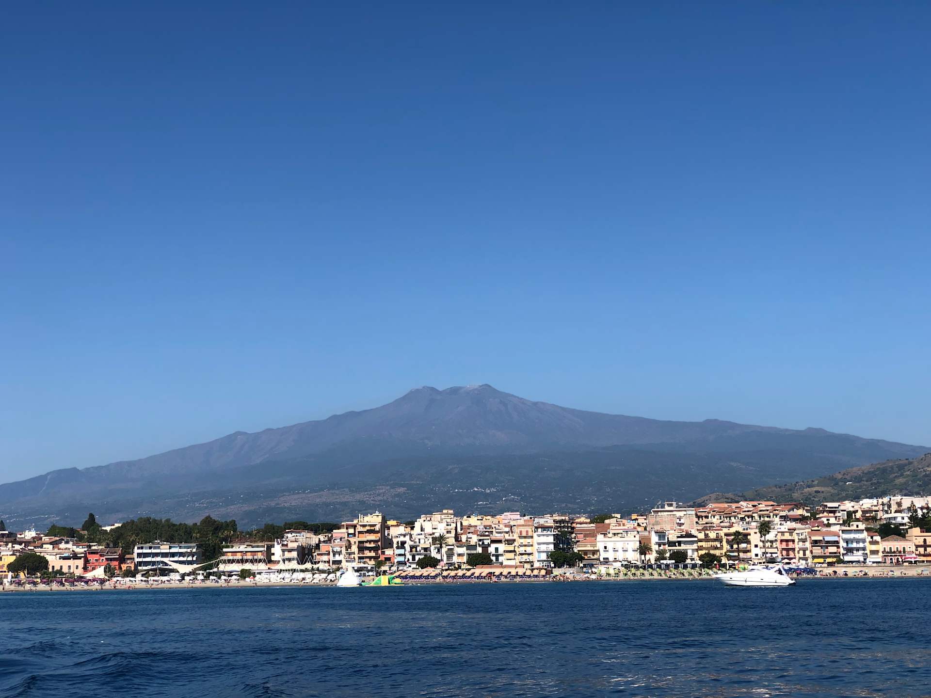 A coastal town nestled along the shore, with a majestic mountain rising in the background against a clear blue sky.