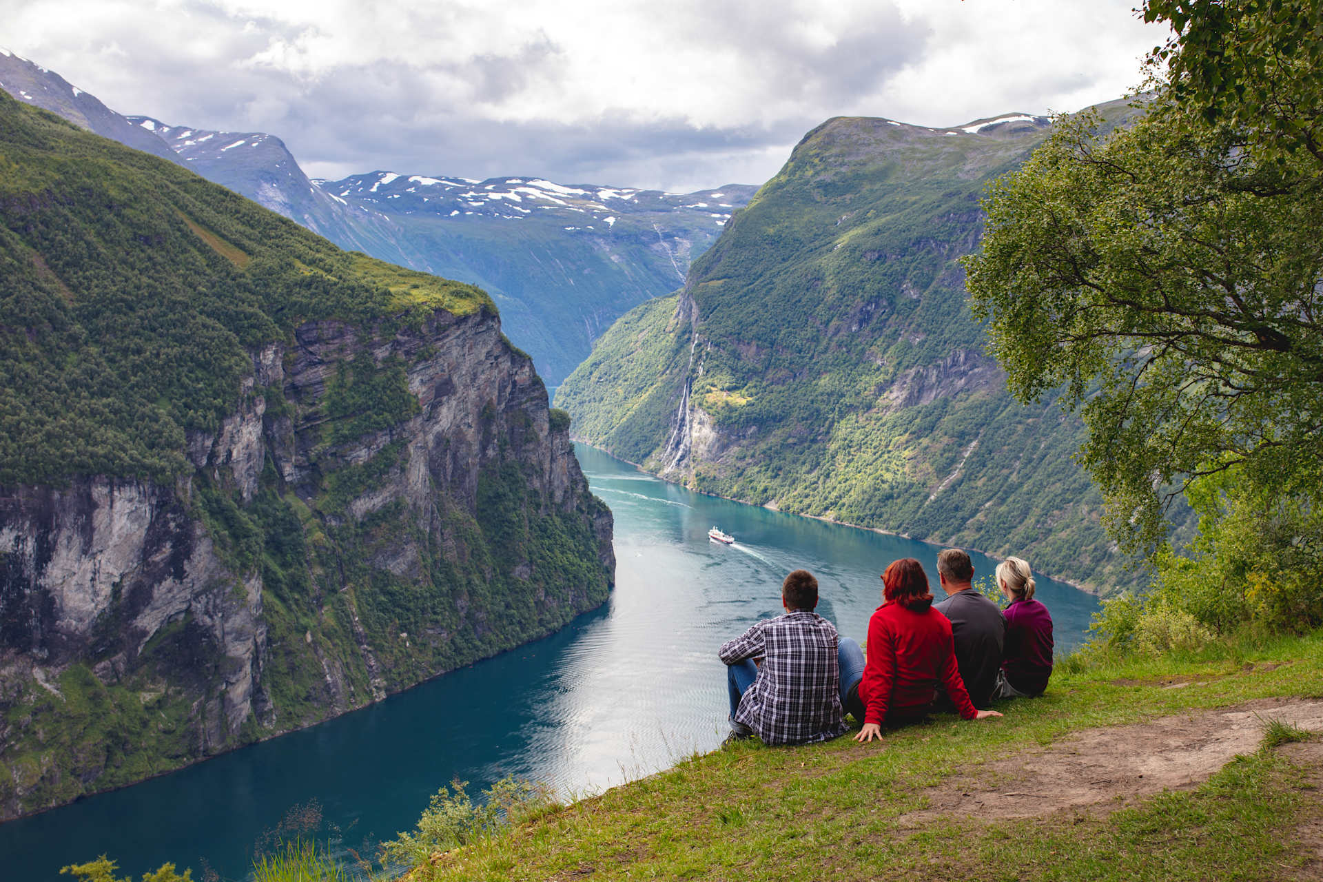 A group of people standing on a grassy hillside overlooking a majestic fjord surrounded by towering mountains in the background.