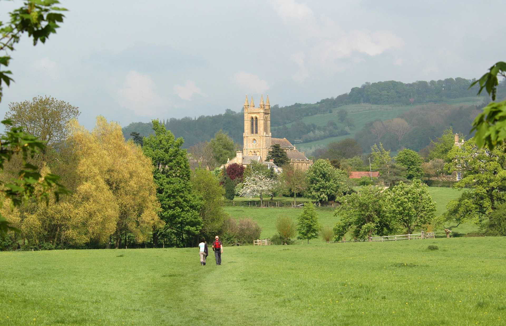 A lush, green meadow in the foreground leads to a picturesque church tower nestled among the rolling hills and trees in the background.