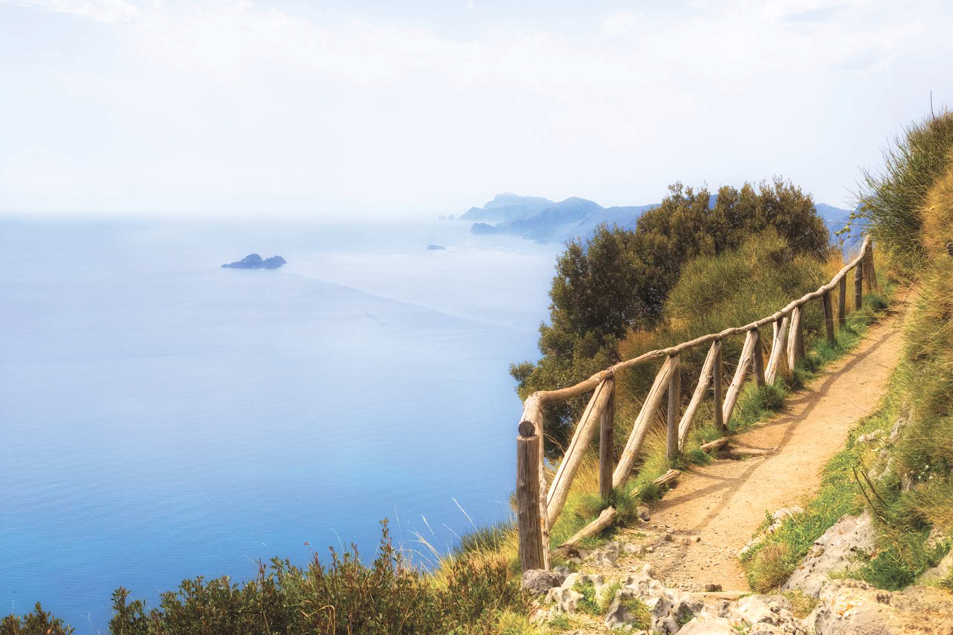 A winding dirt path with a wooden railing runs along the edge of a cliff, overlooking a vast expanse of blue ocean and distant hazy mountains.