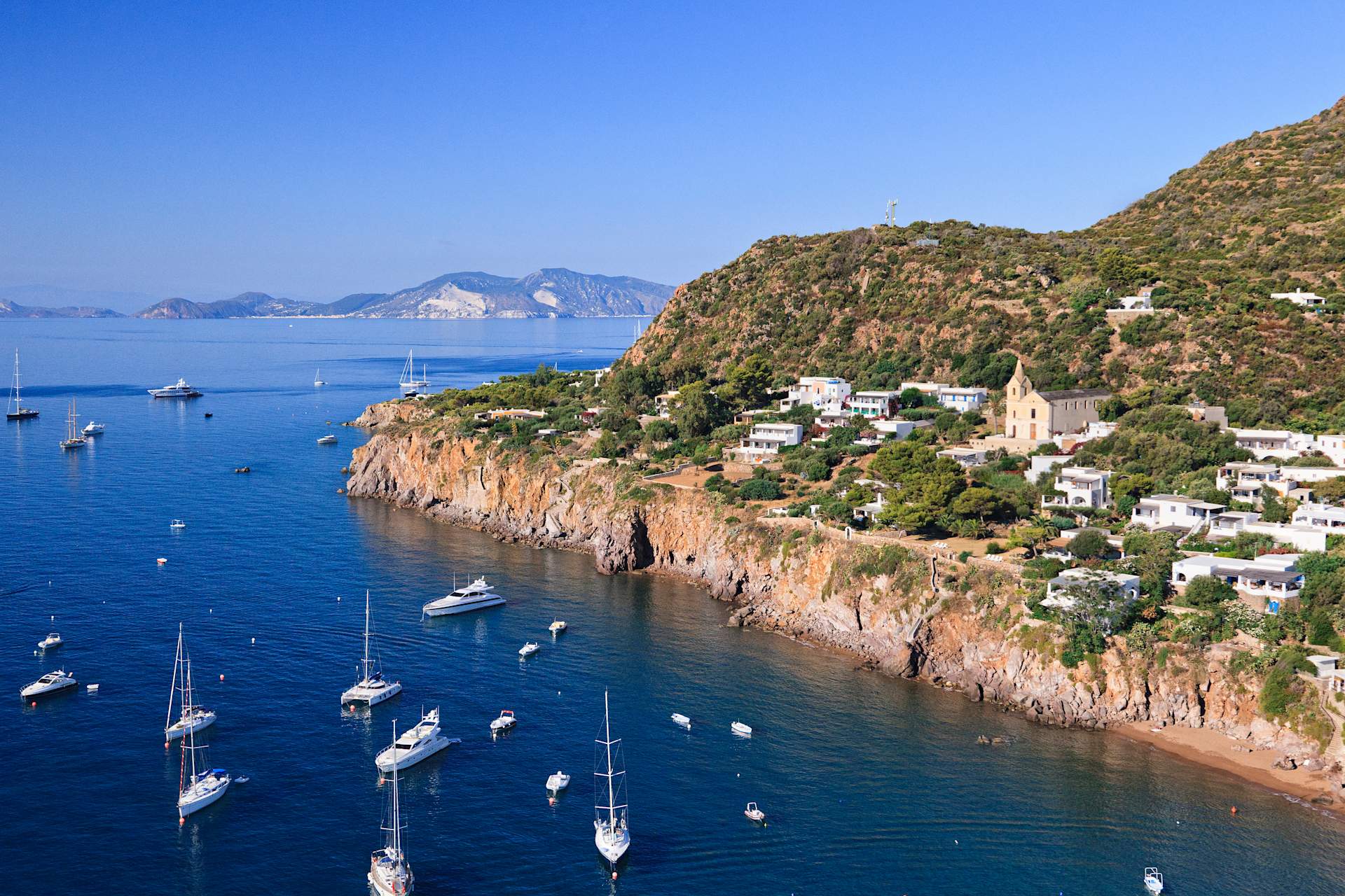 A picturesque coastal town nestled among rugged cliffs, with sailboats and yachts dotting the tranquil turquoise waters in the foreground.
