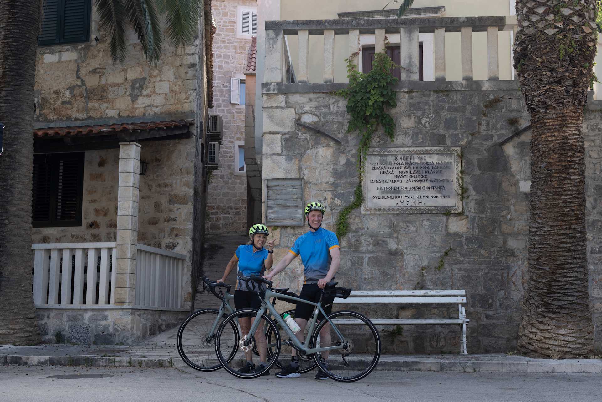 Two cyclists in blue and white jerseys stand with their bicycles in front of a stone building with a plaque on the wall.