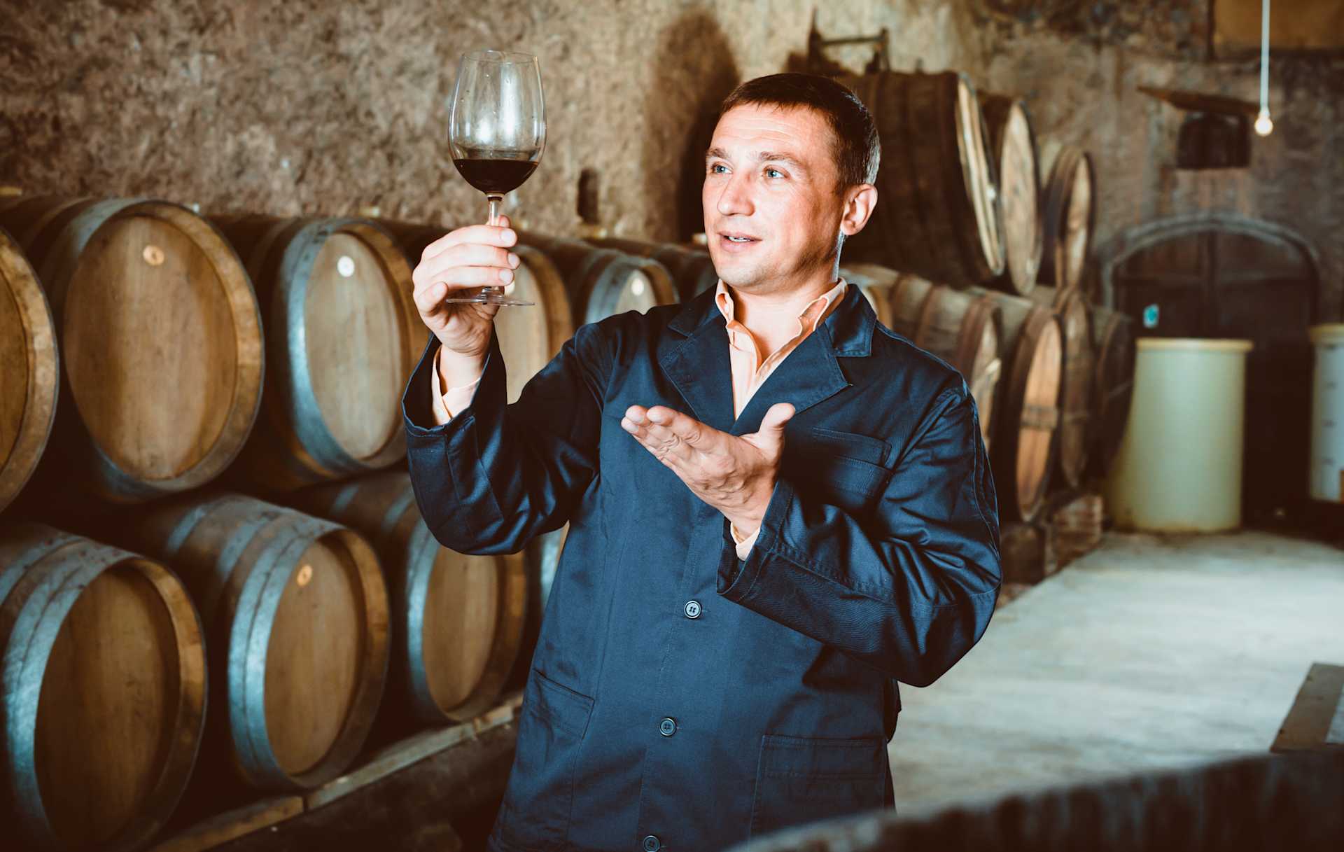 A man in a dark blue jacket stands in a wine cellar, holding a glass of red wine and gesturing as he speaks.