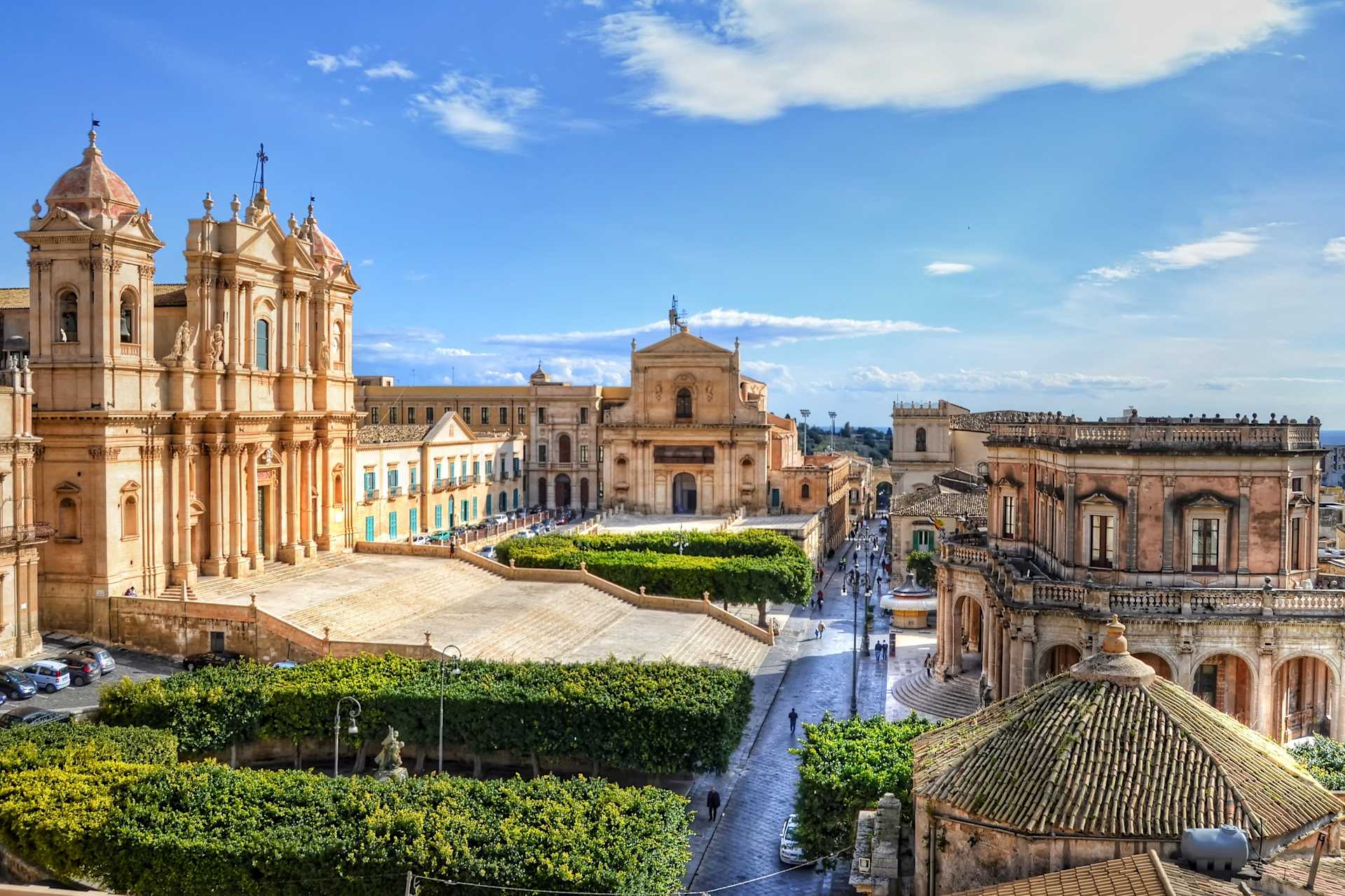A picturesque town square with ornate, historic buildings, lush greenery, and a winding path leading through the scene, set against a bright blue sky with fluffy clouds.