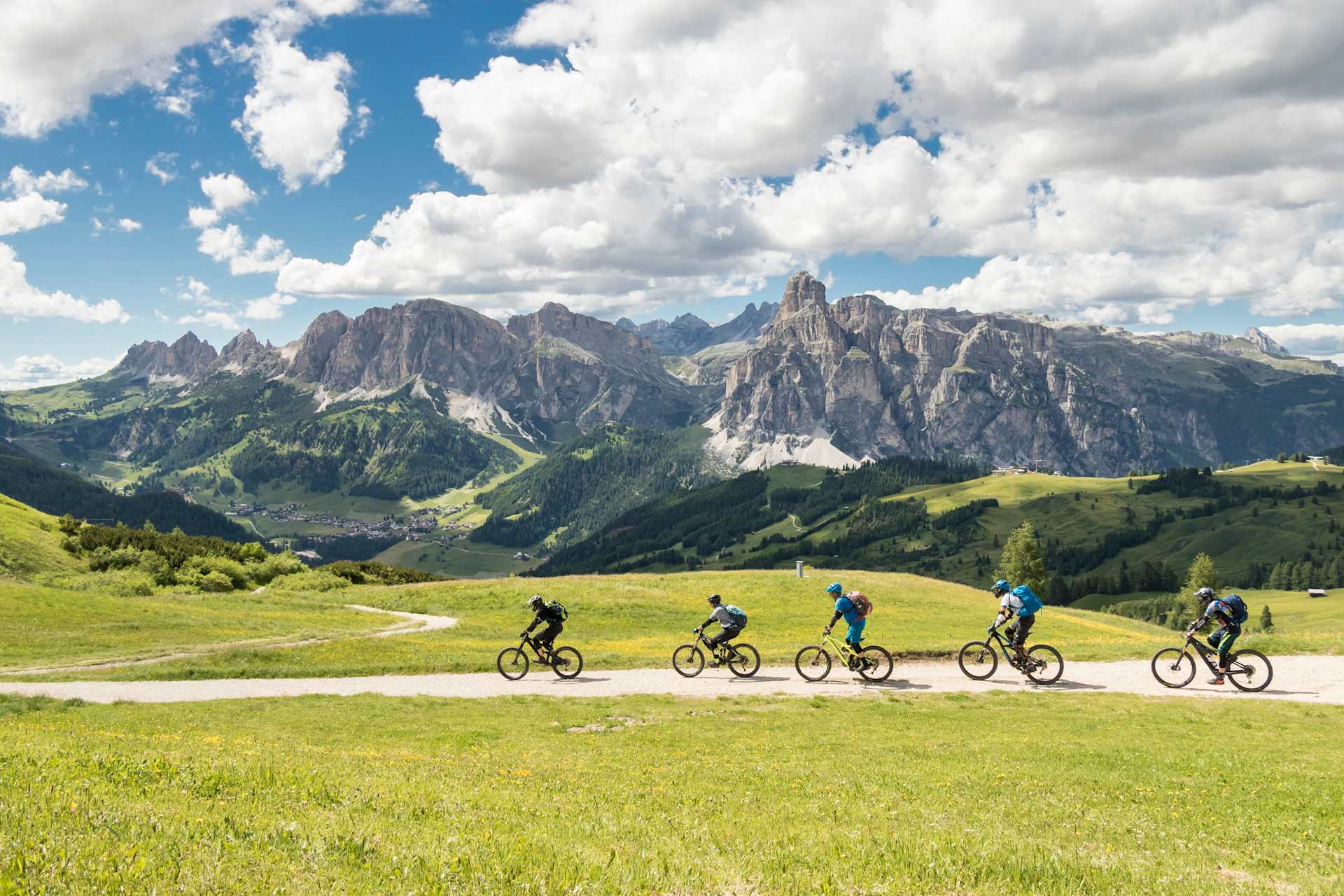A group of cyclists riding through a picturesque mountain landscape, with towering peaks and lush green meadows in the background.