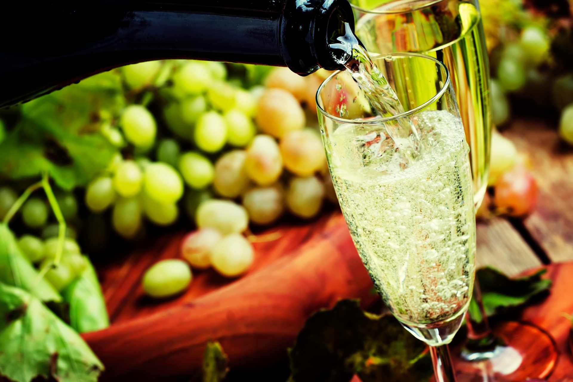 A glass of sparkling wine being poured against a backdrop of fresh green grapes and vibrant red fabric.