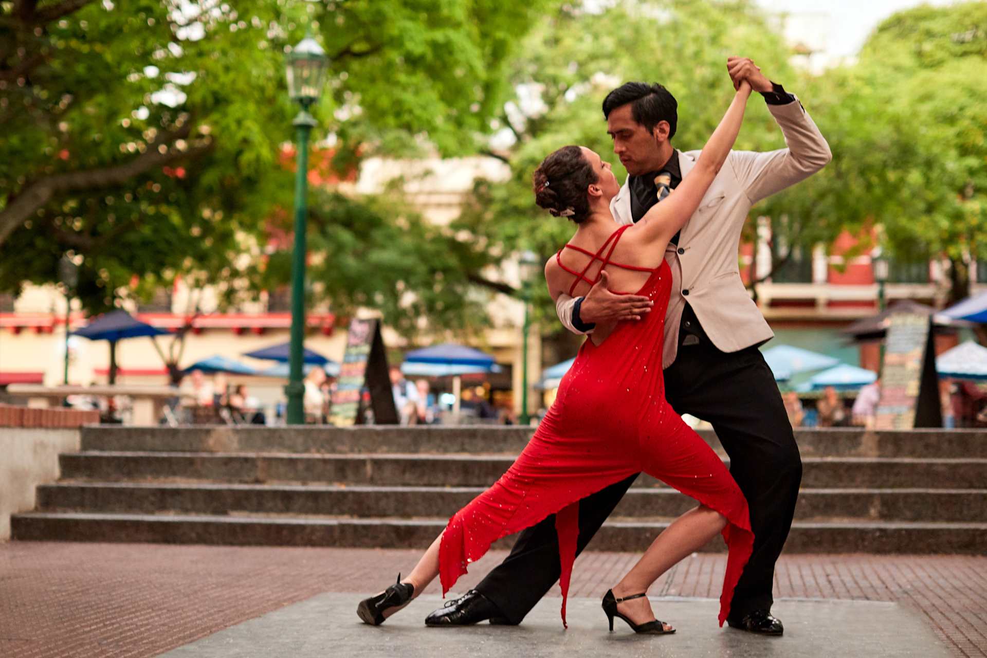 A couple dressed in formal attire, the woman in a vibrant red dress and the man in a suit, are engaged in a passionate dance on a set of stairs surrounded by lush greenery and urban scenery.