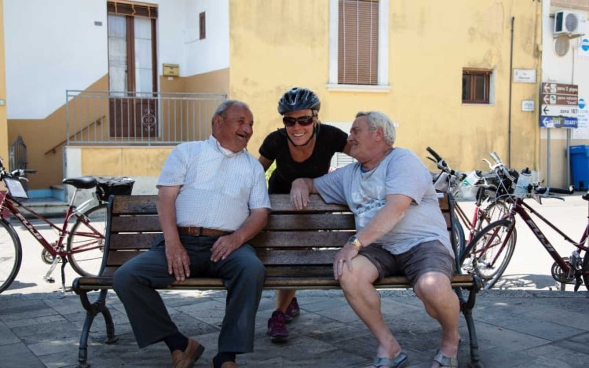 Three elderly individuals, two men and one woman, are seated on a bench in an outdoor setting with bicycles and buildings visible in the background.