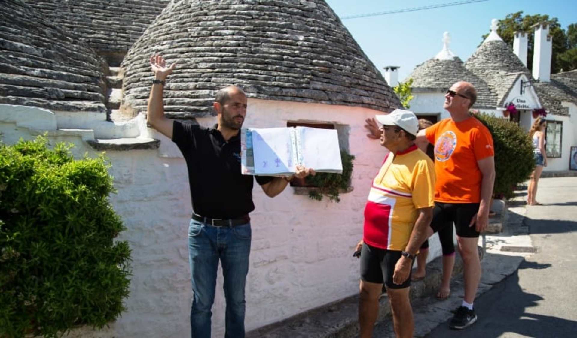 The image shows a group of people standing in front of traditional cone-shaped buildings, likely in a rural or small-town setting, with one person holding up what appears to be a painting or drawing.