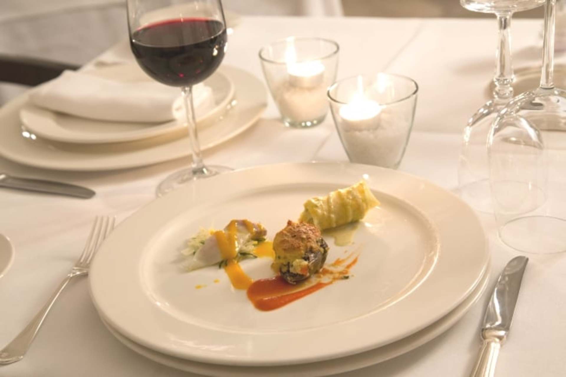 A white plate with a dish featuring a cooked item, likely meat, accompanied by a glass of red wine and other tableware on a table setting.