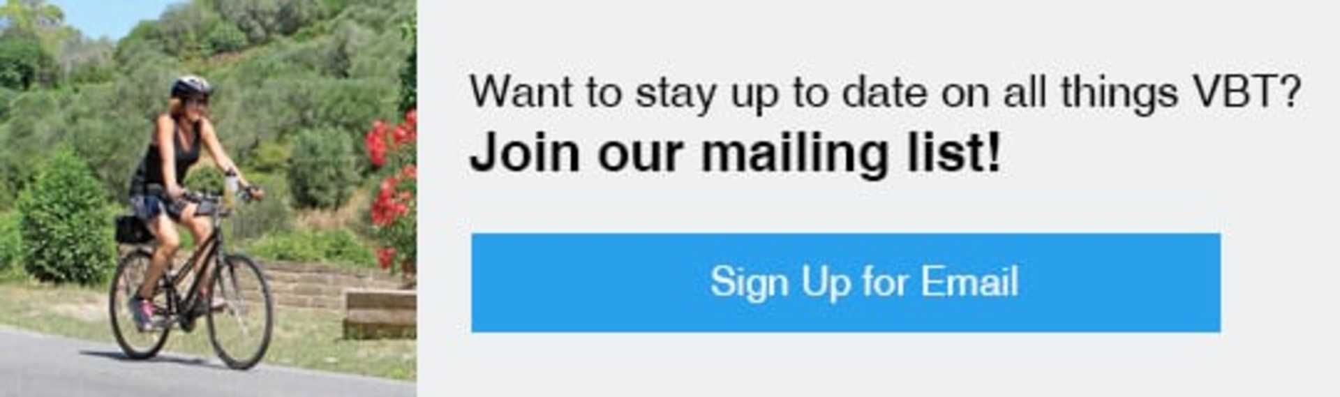 The image shows a sign inviting viewers to join a mailing list in order to stay up to date on all the latest information. The background features a lush, green landscape with some red flowers visible.