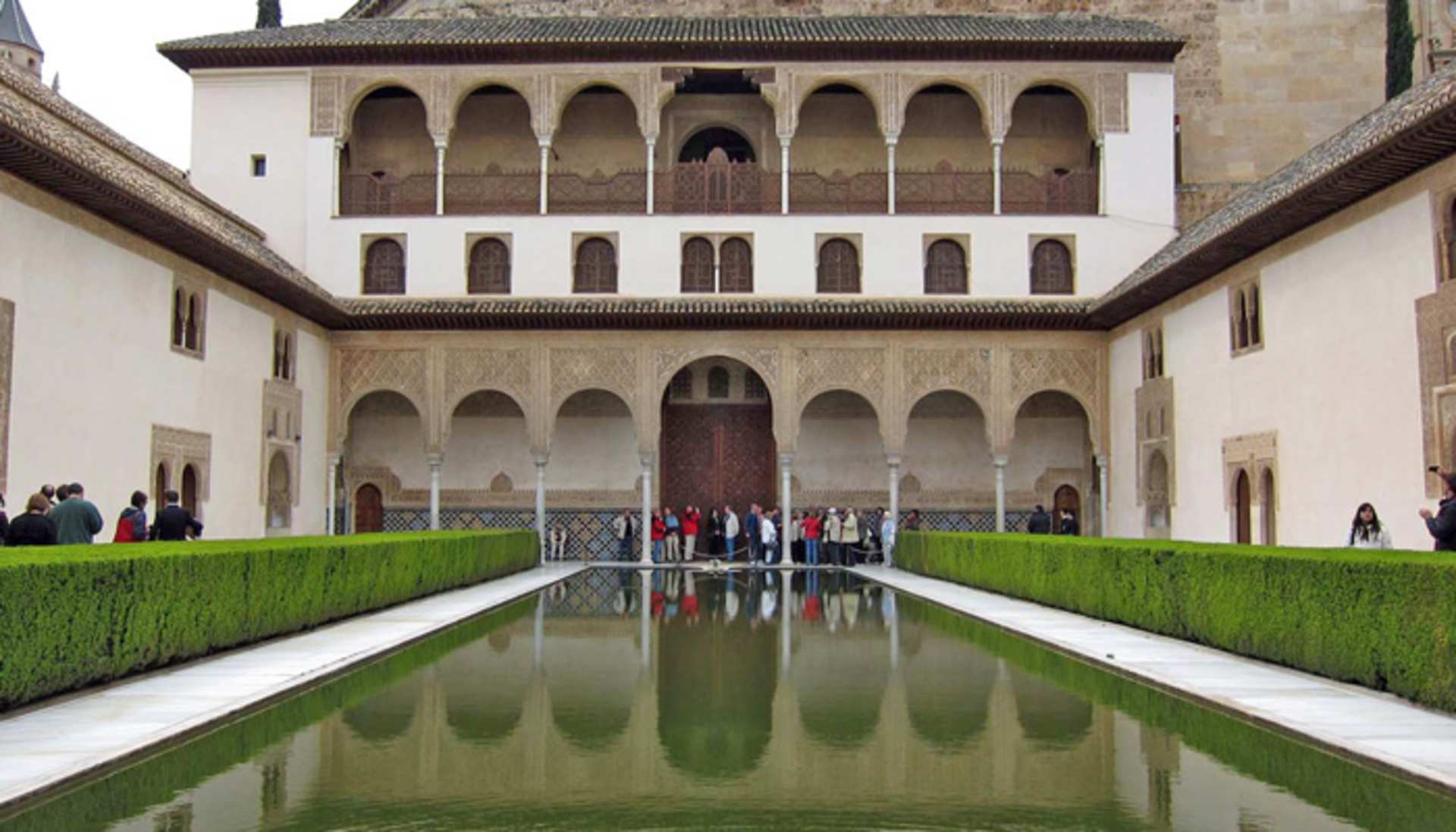 A grand, ornate building with arched windows and a central tower is reflected in a tranquil pool of water, surrounded by lush green grass in the foreground.