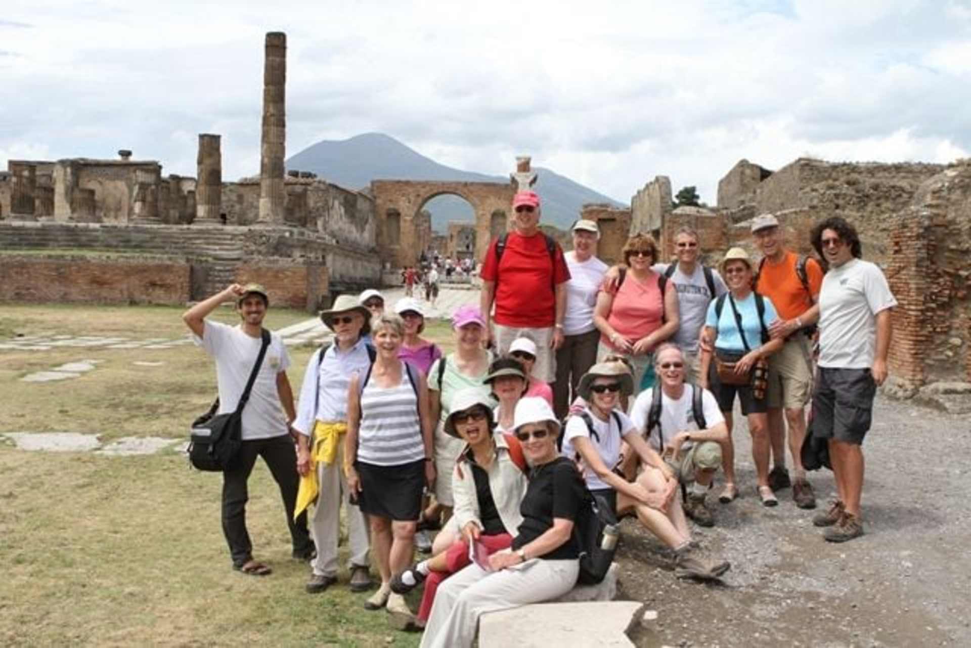 A group of people, dressed in casual attire, are gathered in the foreground of an ancient archaeological site with ruins and a mountain backdrop.