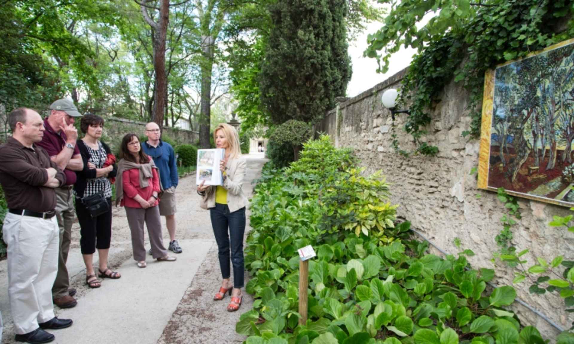 A group of people standing on a path surrounded by lush greenery, including trees, vines, and other vegetation, with a stone wall visible in the background.