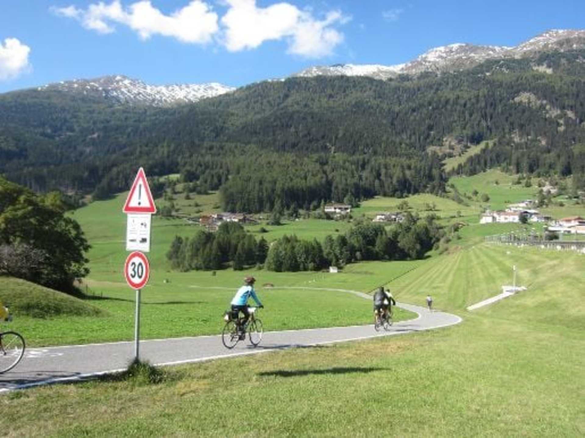 A scenic mountain landscape with cyclists riding on a winding road, surrounded by lush green forests and snow-capped peaks in the background.