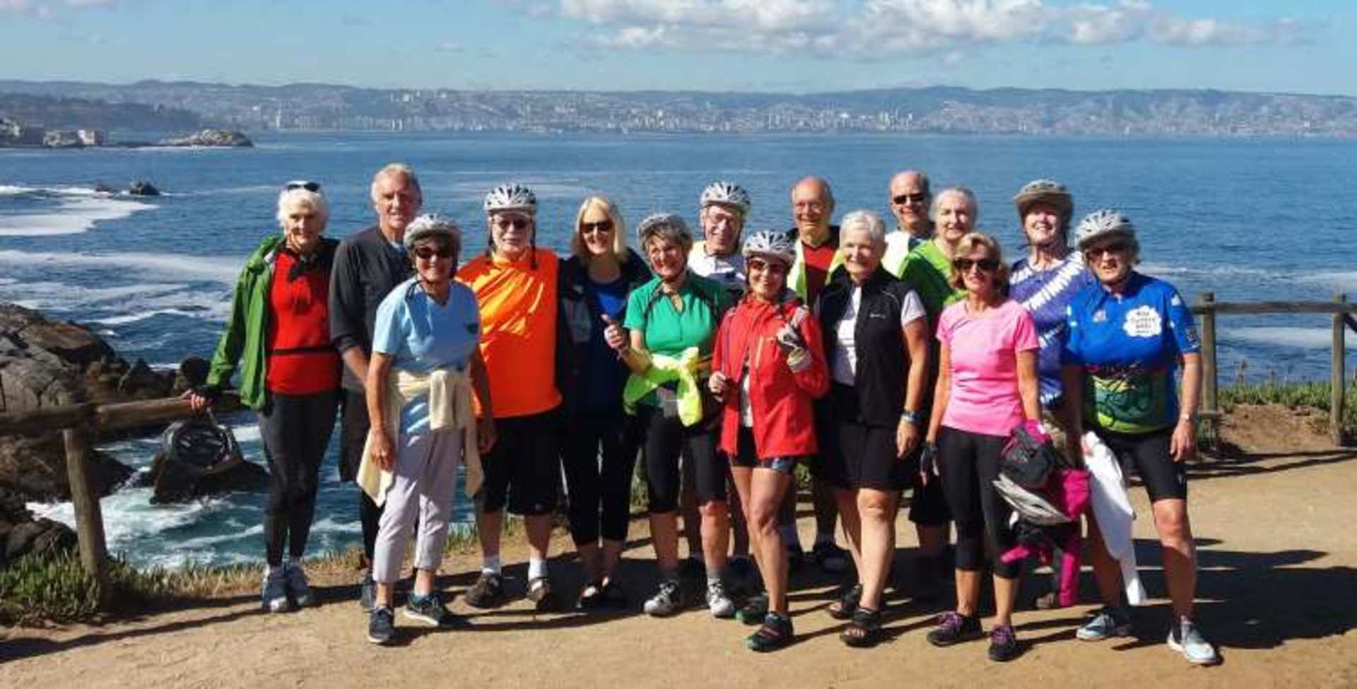 A group of people in colorful athletic wear standing together on a coastal path overlooking a scenic ocean view with mountains in the background.