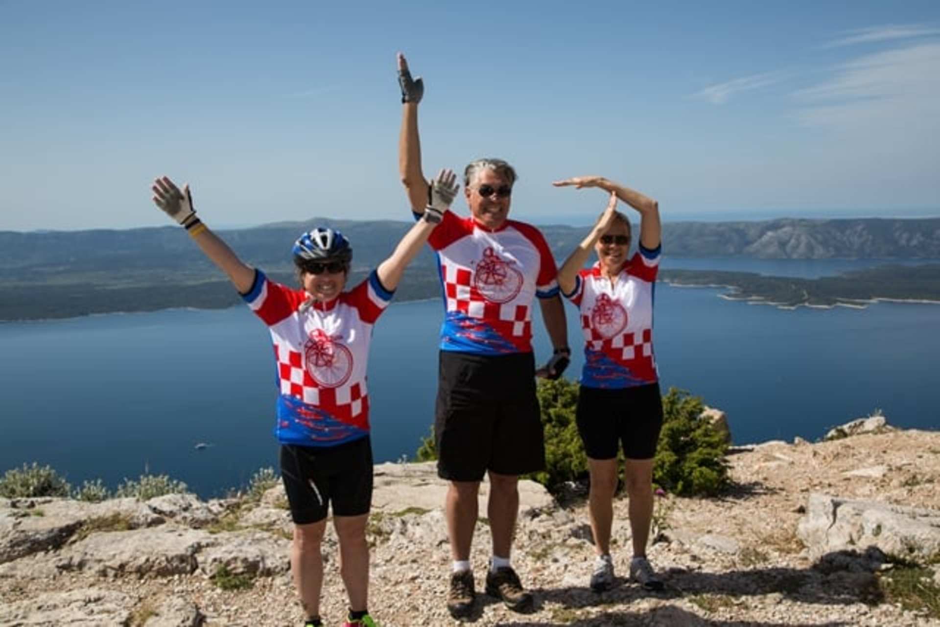 Three people in athletic clothing stand on a rocky outcrop overlooking a scenic body of water surrounded by mountains in the background.