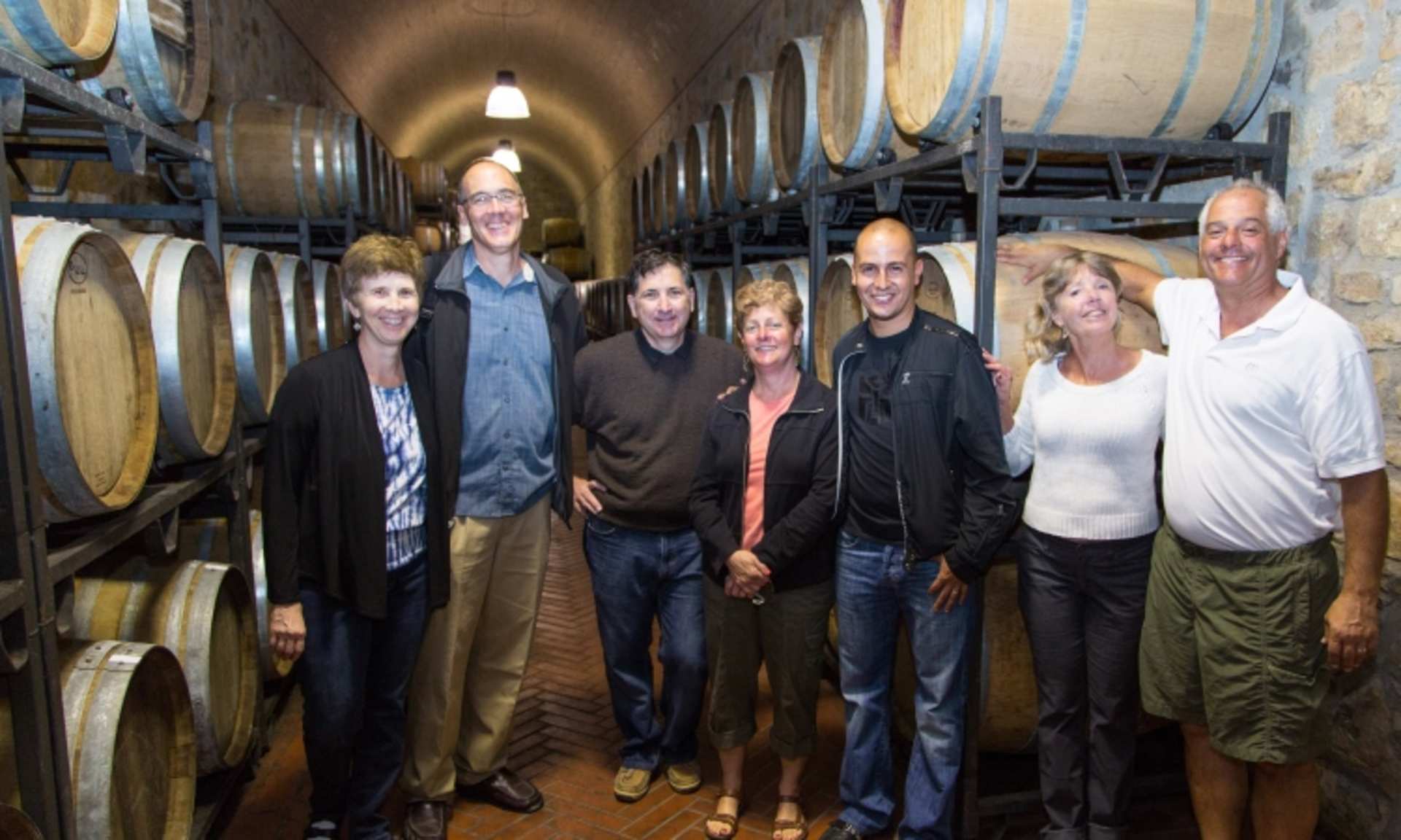 A group of people standing in a wine cellar surrounded by rows of oak barrels.