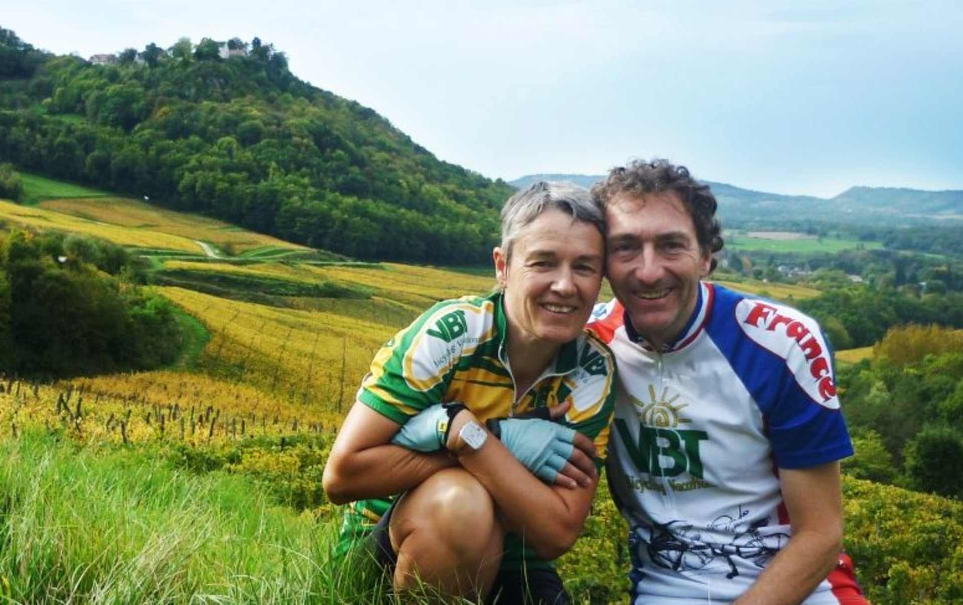 The image shows a middle-aged couple embracing in a scenic, mountainous landscape with lush green vegetation and rolling hills in the background.