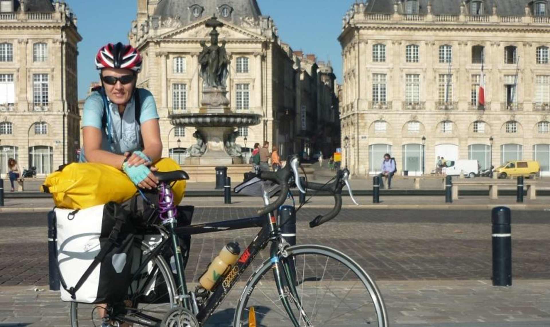 A cyclist wearing a helmet and bright clothing stands in the foreground, with a historic, ornate building and plaza visible in the background.