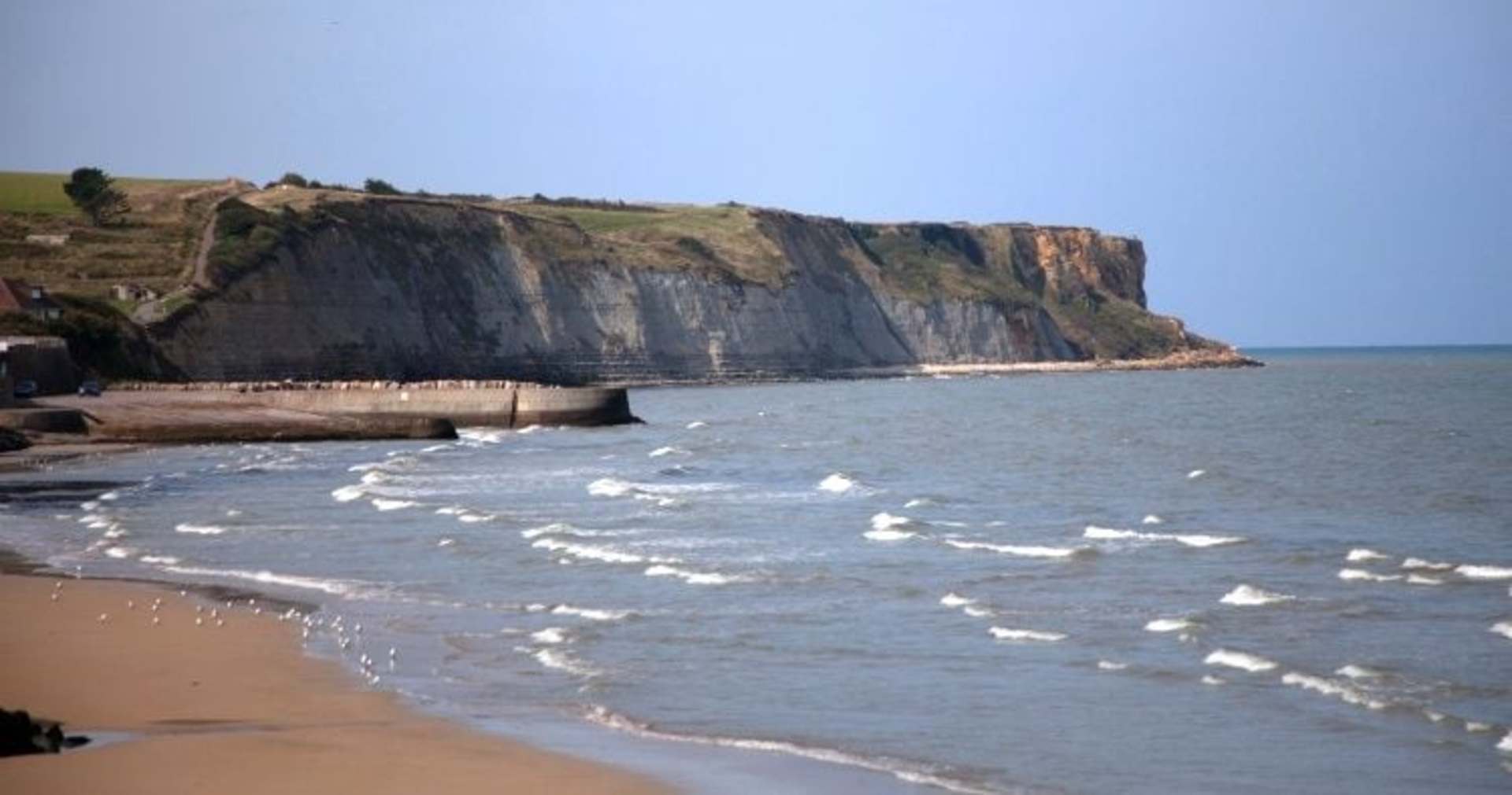 A rugged, rocky coastline with a sandy beach in the foreground, overlooking the calm, blue waters of the ocean under a clear sky.