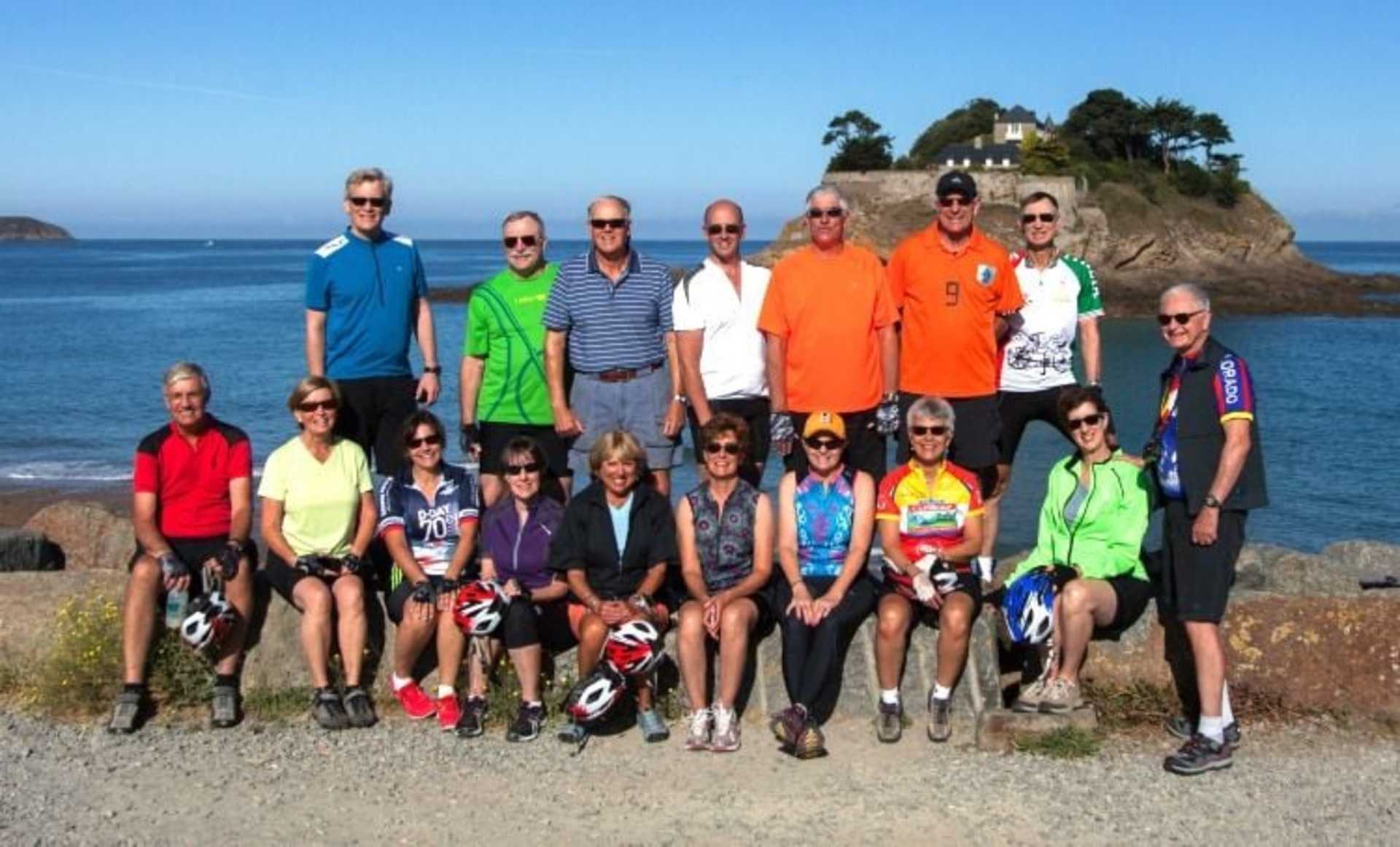 A group of people, dressed in various colorful outfits, standing on a beach with a rocky cliff and ocean in the background.