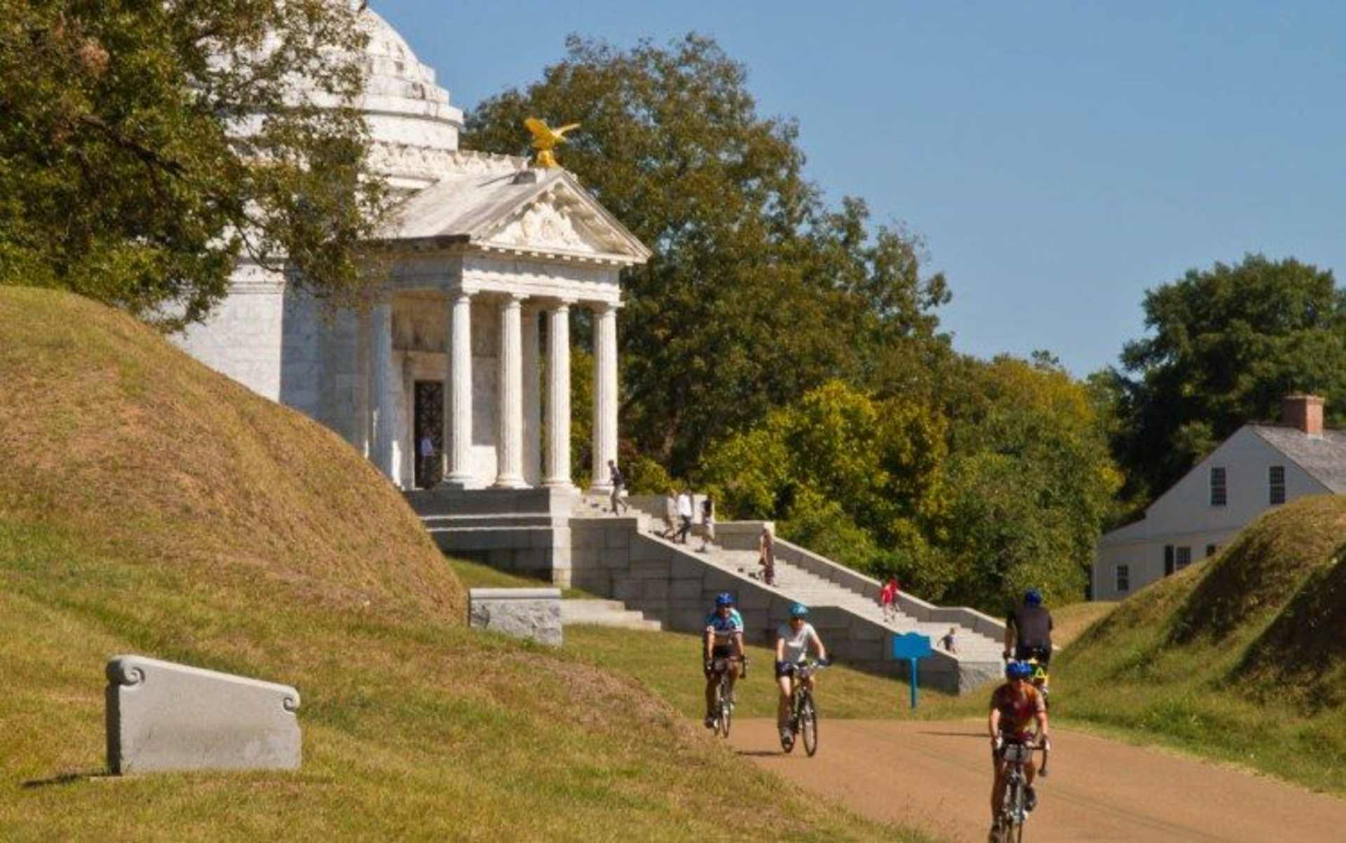 A group of cyclists riding on a dirt path in front of a grand, columned building surrounded by lush greenery and a clear blue sky.