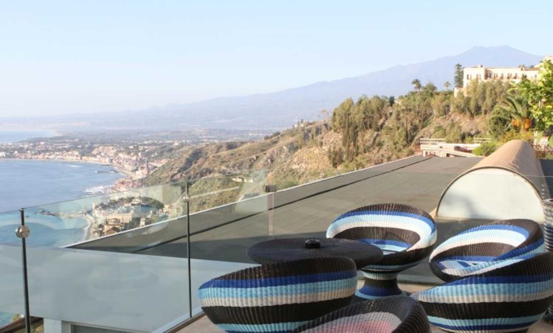 A glass-enclosed balcony overlooking a scenic coastal town, with striped sandals and a glass table in the foreground.