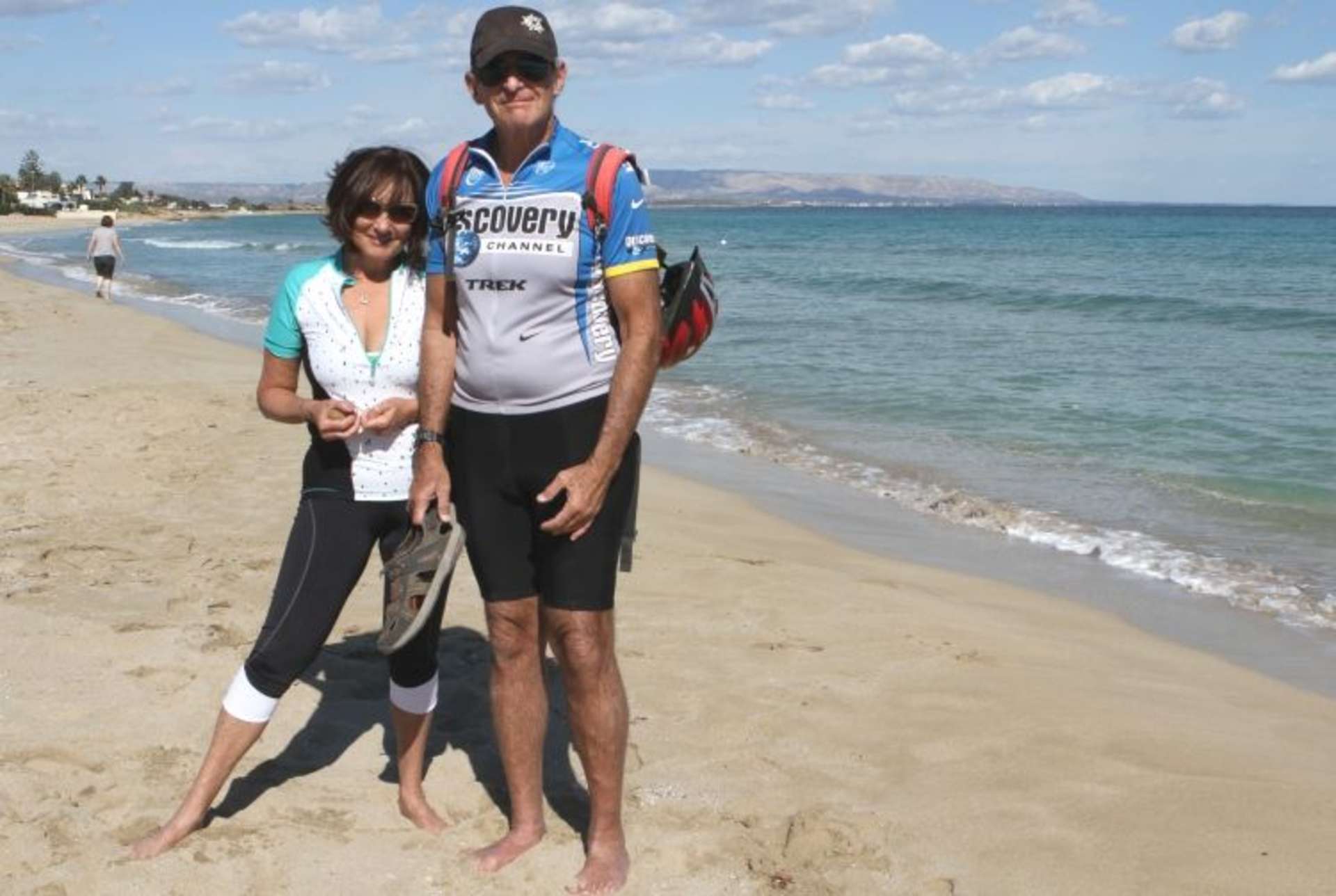 A couple standing on a sandy beach, with the ocean and mountains visible in the background.
