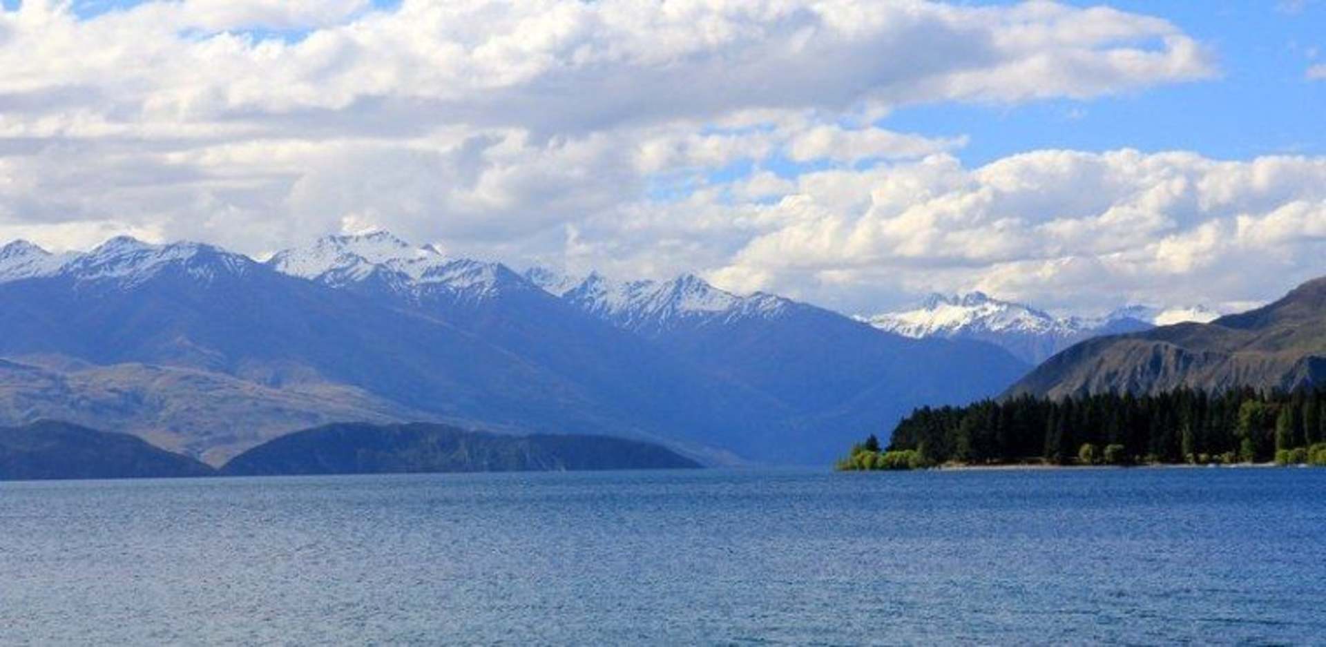 A serene lake surrounded by majestic snow-capped mountains and fluffy white clouds in the blue sky.
