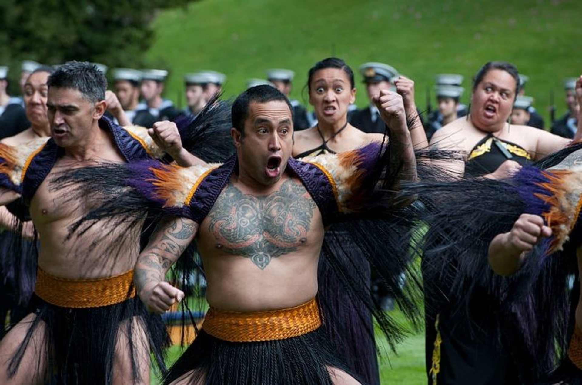 A group of people wearing colorful, feathered costumes and striking dramatic poses in an outdoor setting with a grassy background.