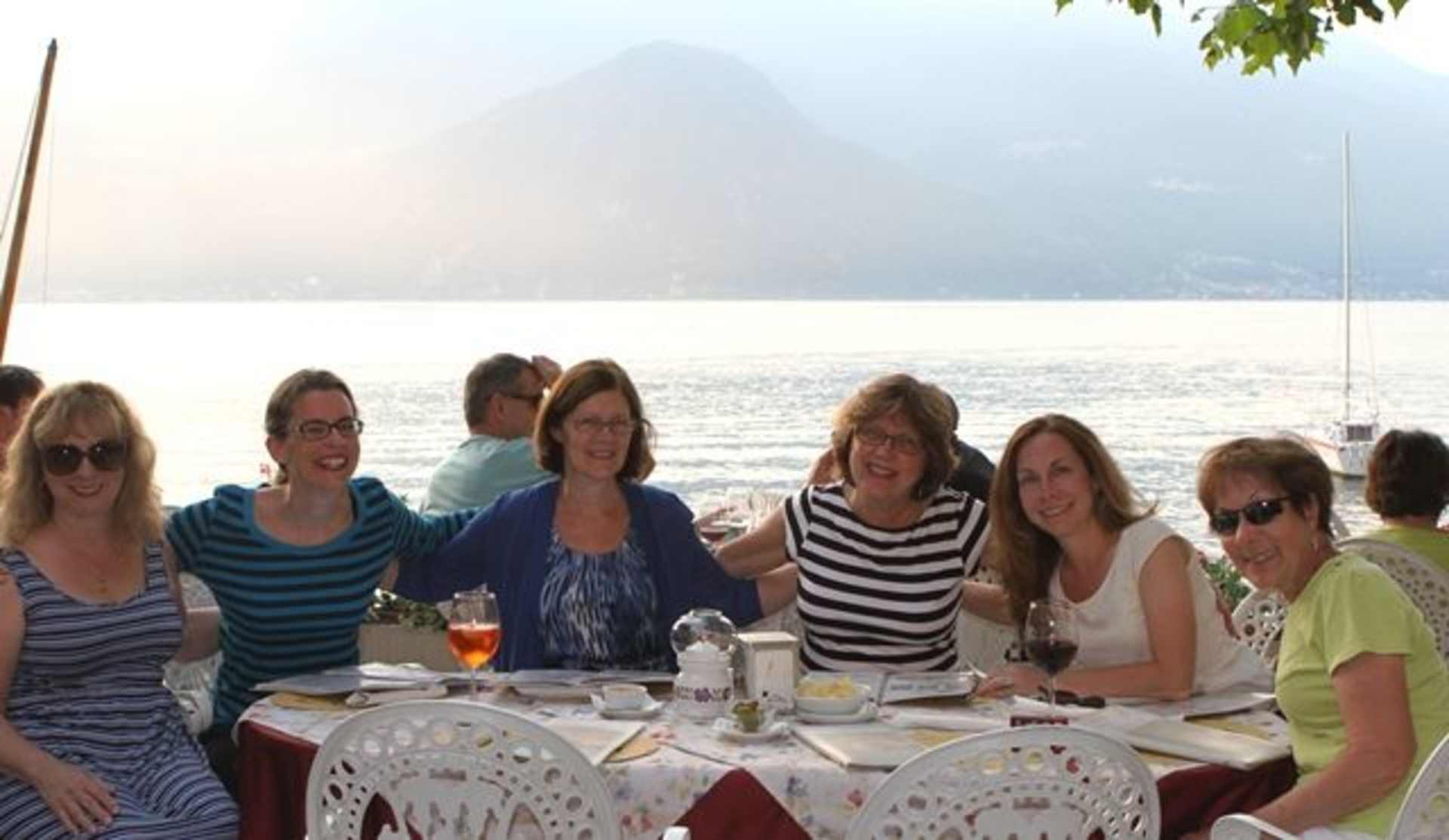 A group of people enjoying a meal outdoors, with a scenic lake and mountains visible in the background.