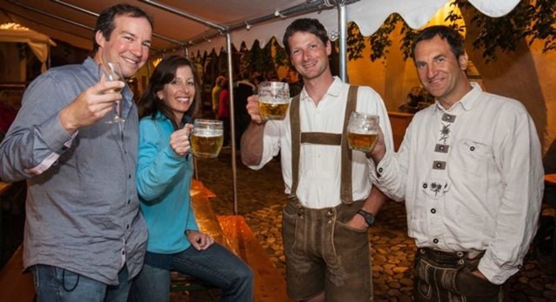A group of four people, three men and one woman, are standing together and holding beer mugs, appearing to be enjoying themselves in a festive, rustic-looking setting with wooden walls and decorations.