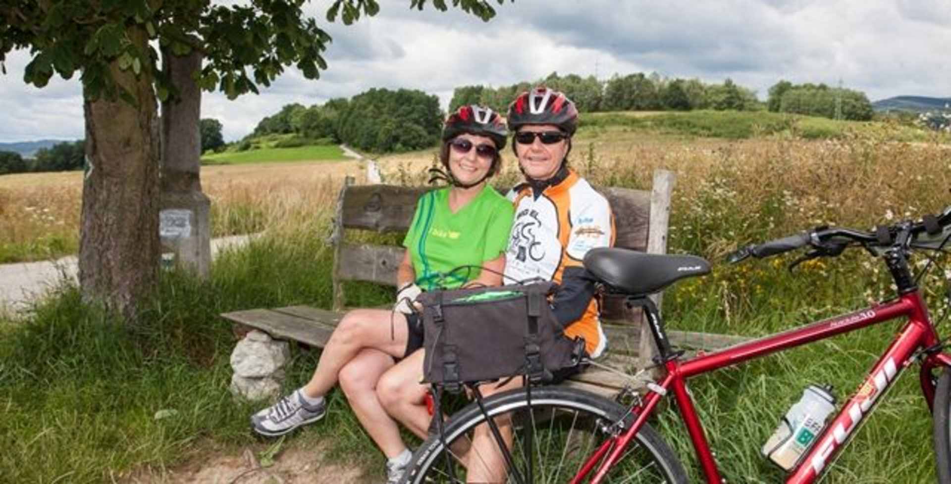 The image shows a couple sitting on a bench in a grassy field, surrounded by trees and mountains in the background. They are wearing cycling gear and appear to be taking a break from their bike ride.