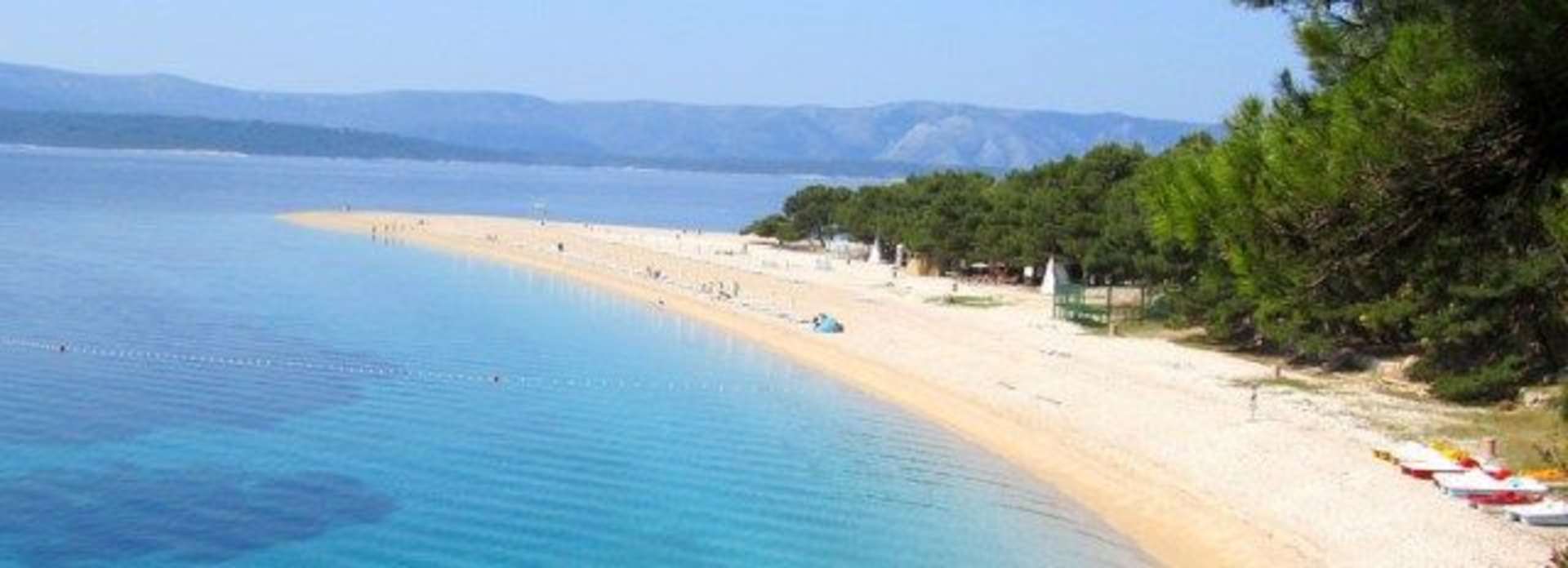 A pristine sandy beach stretches along the turquoise waters of a scenic lake, surrounded by lush pine trees and towering mountains in the distance.