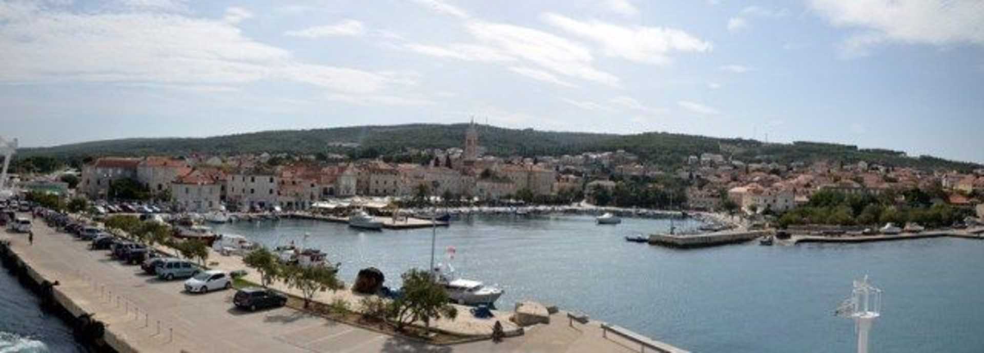 A picturesque coastal town with colorful buildings, boats in the harbor, and a hilly landscape in the background.