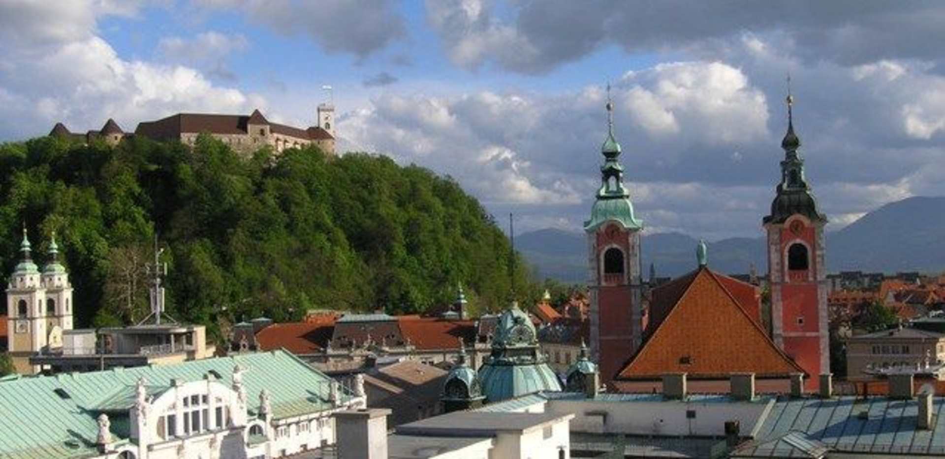 A picturesque European city skyline with ornate church spires and rooftops surrounded by lush greenery against a dramatic cloudy sky.