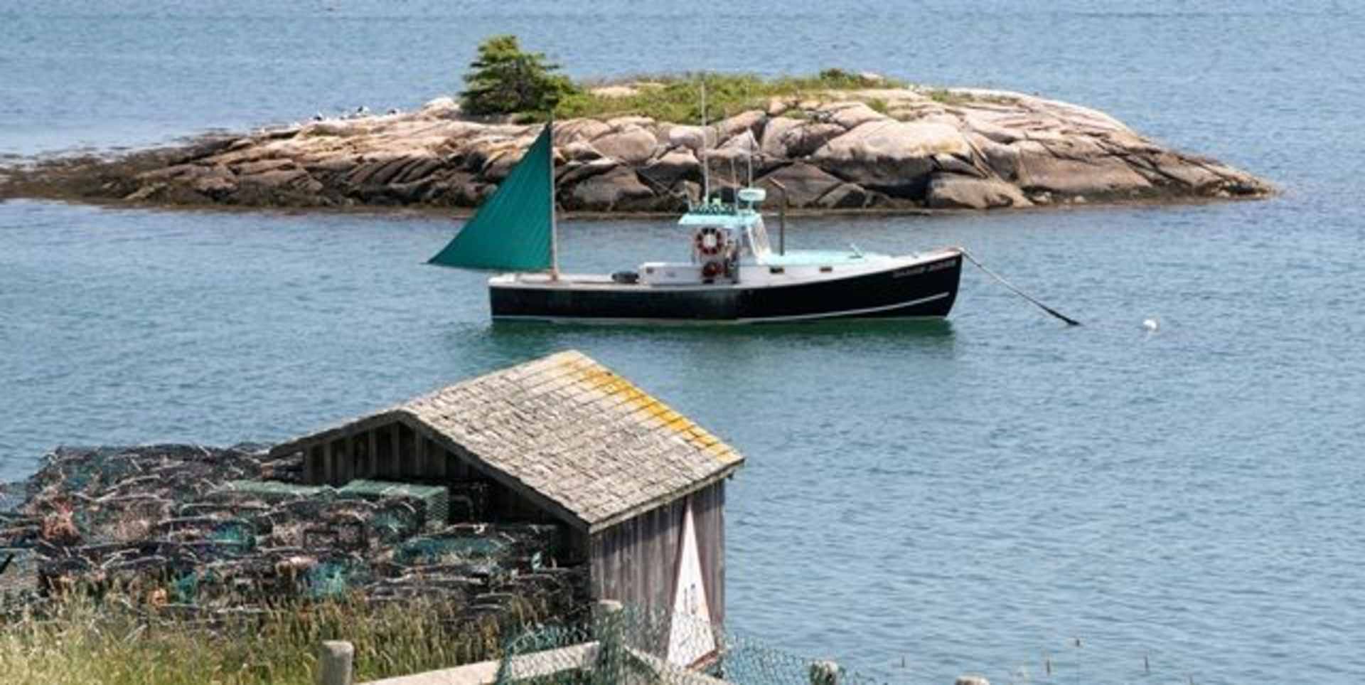 A small green boat with a sail is anchored near a rocky island, with a wooden structure in the foreground overlooking the calm, blue waters.