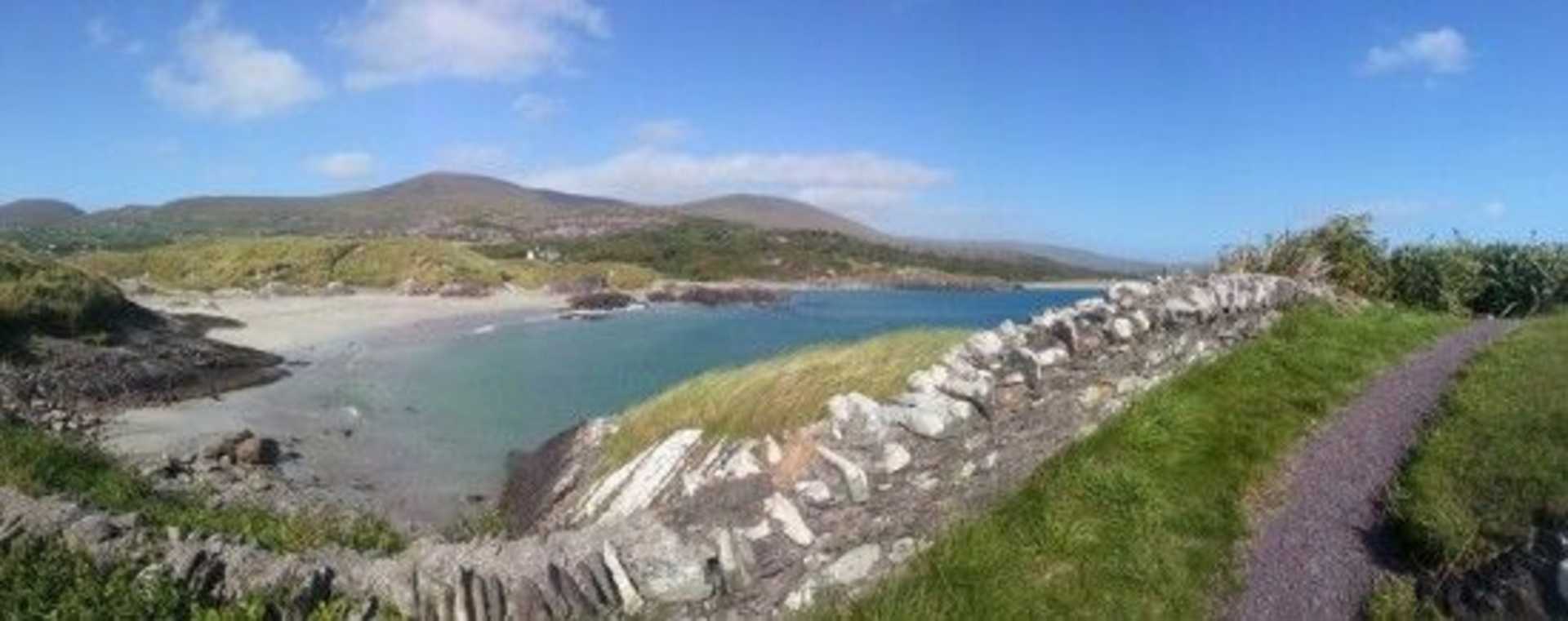 A scenic coastal landscape with a rocky shoreline, lush green hills, and a tranquil bay surrounded by mountains in the background under a clear blue sky.