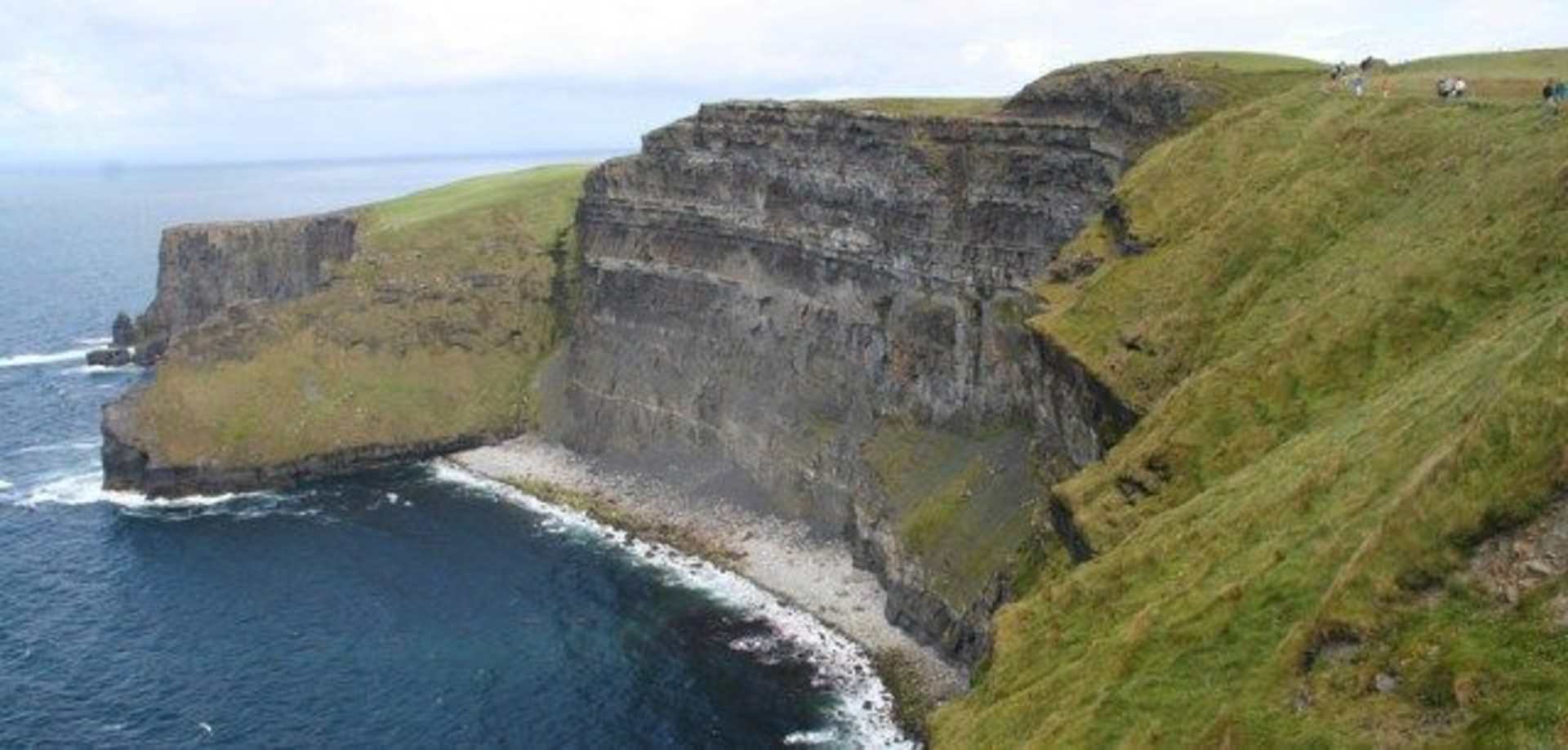 Rugged cliffs with a grassy top overlooking the vast expanse of the ocean, with a few people visible on the cliff's edge.