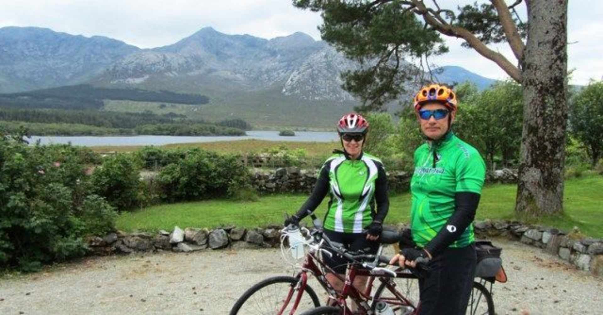 Two cyclists in cycling gear stand on a dirt path, with a scenic mountain landscape visible in the background.
