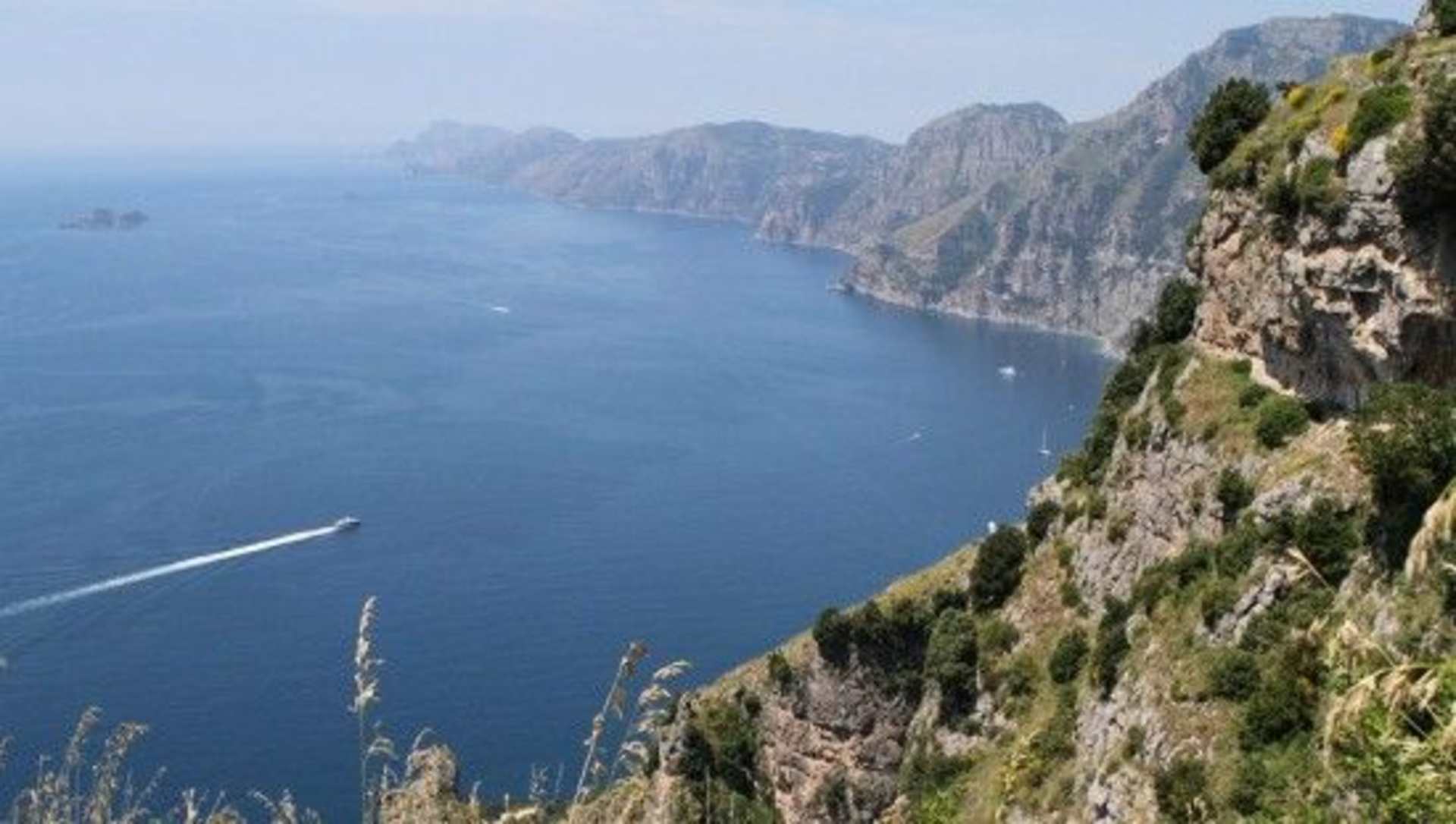 Rugged cliffs plunge into the deep blue waters of the sea, with small boats dotting the horizon against a backdrop of towering mountains.