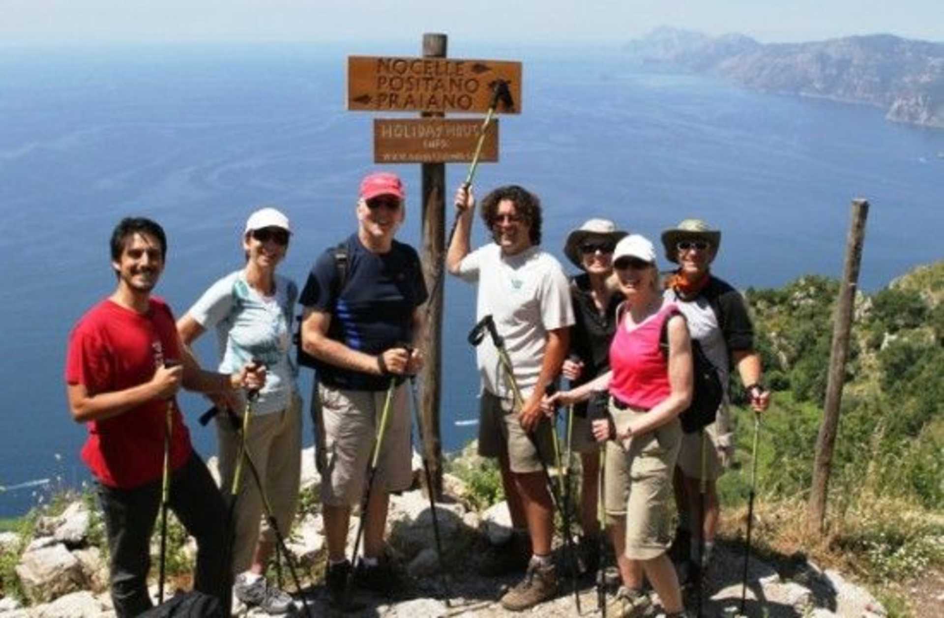 A group of hikers standing in front of a signpost on a scenic trail overlooking a vast body of water and mountainous terrain in the background.