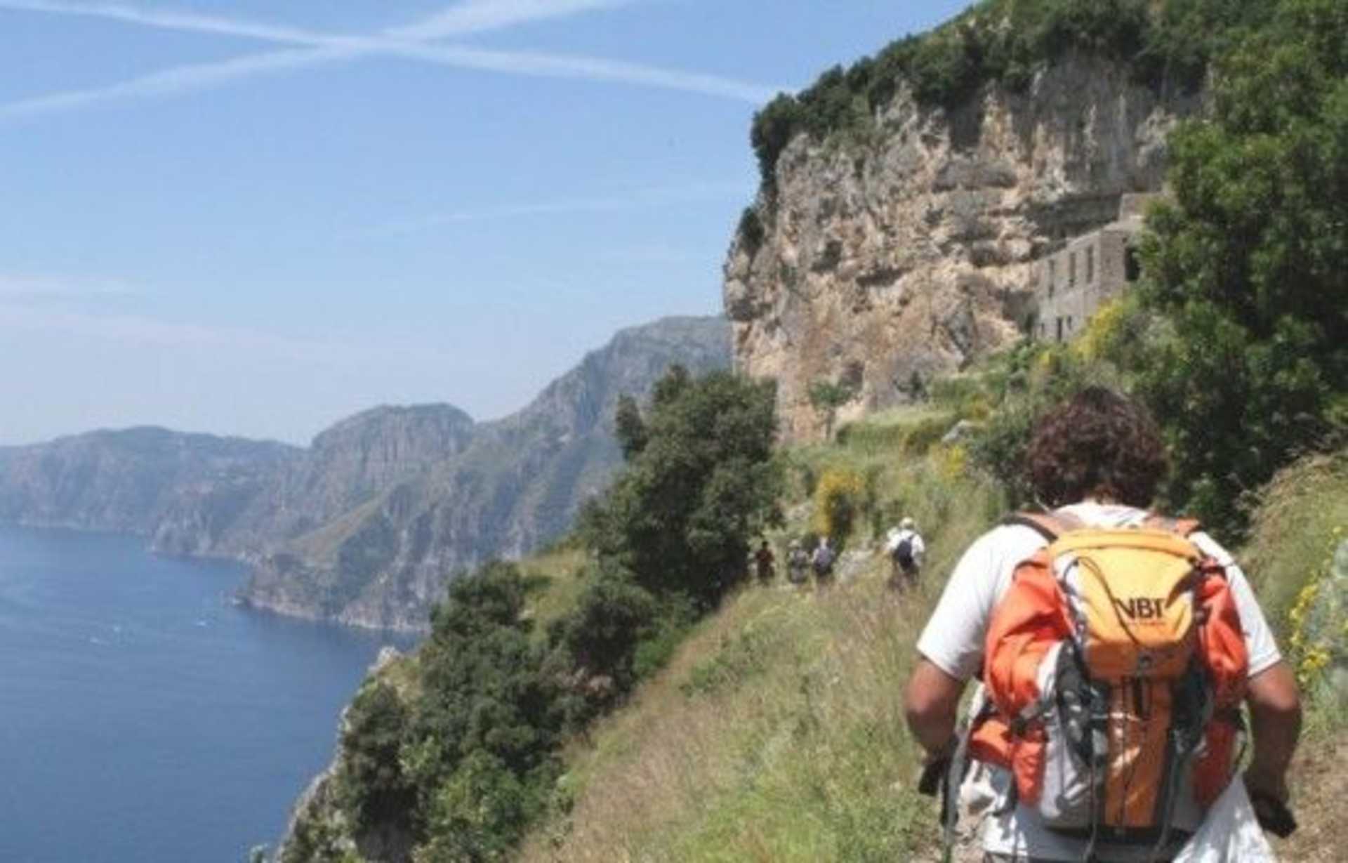 A person wearing a backpack stands on a hiking trail overlooking a scenic coastal landscape with steep cliffs and a body of water in the background.