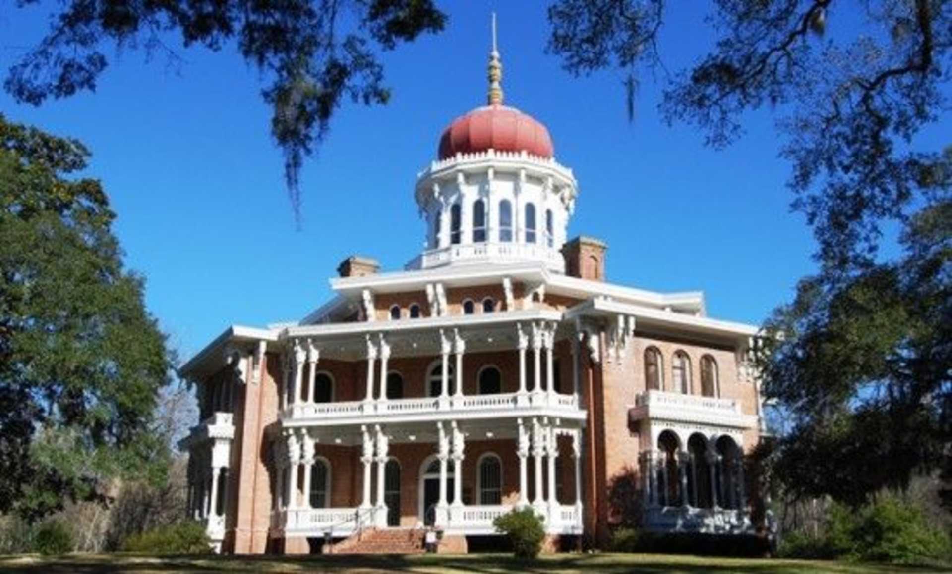 A grand, ornate building with a distinctive red-domed roof stands amidst lush greenery, framed by the clear blue sky above.
