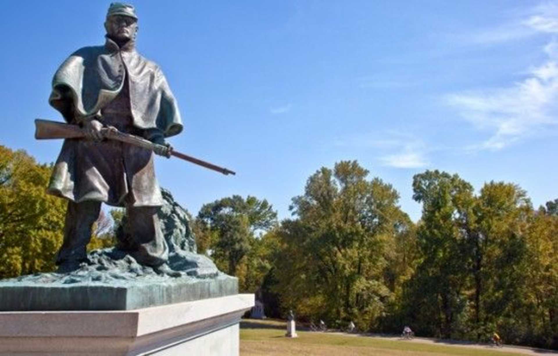 A bronze statue of a soldier stands on a pedestal, surrounded by a lush, autumnal landscape with trees in the background.