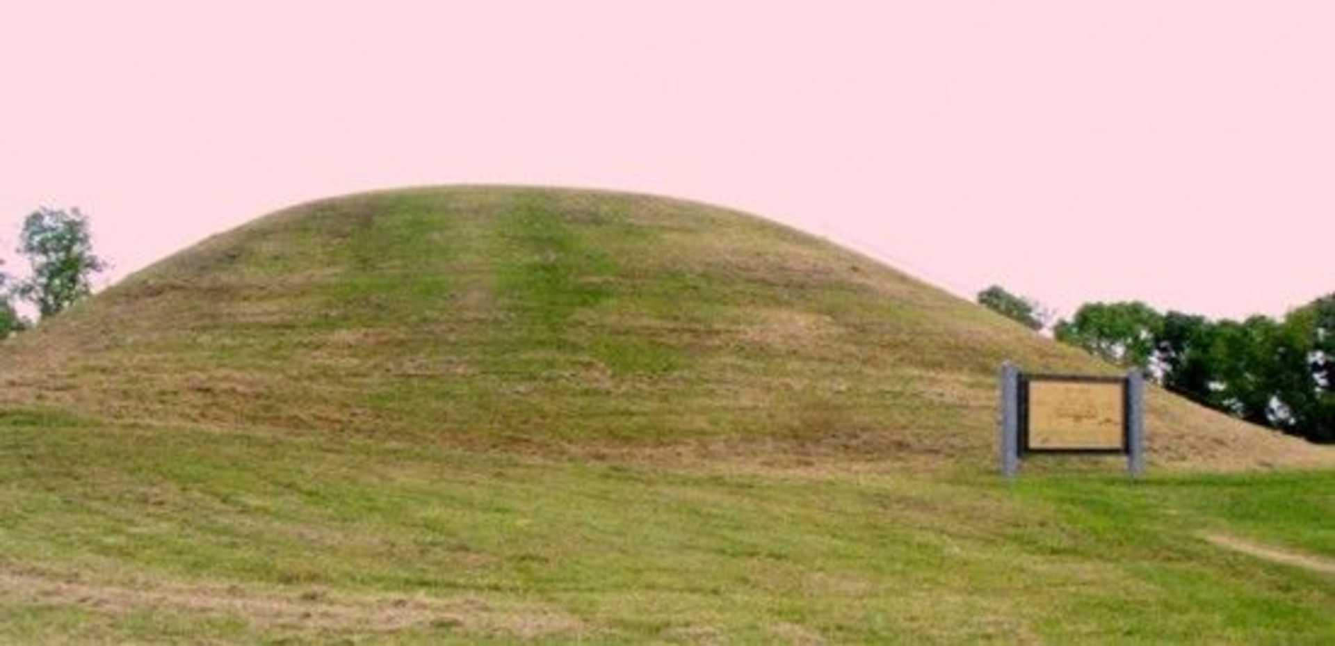 A large, grassy mound with a sign on it stands in a landscape with trees in the background.