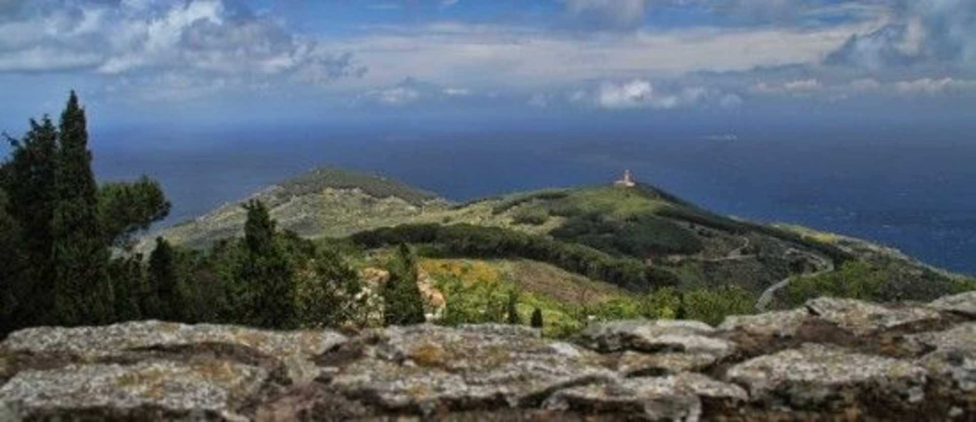 A rugged, mountainous landscape with a winding path leading up to a small structure atop a hill, set against a backdrop of a vast, blue ocean and cloudy sky.