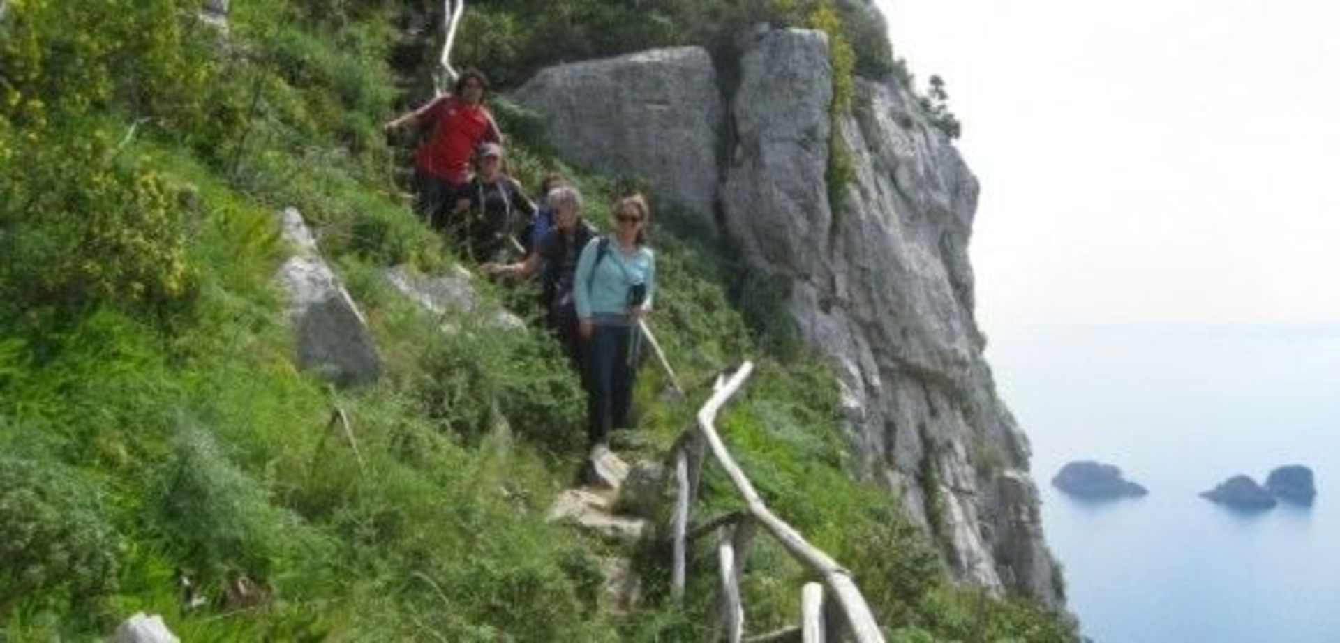 Two people are rock climbing on a steep, moss-covered cliff face, with a serene lake and mountains visible in the background.